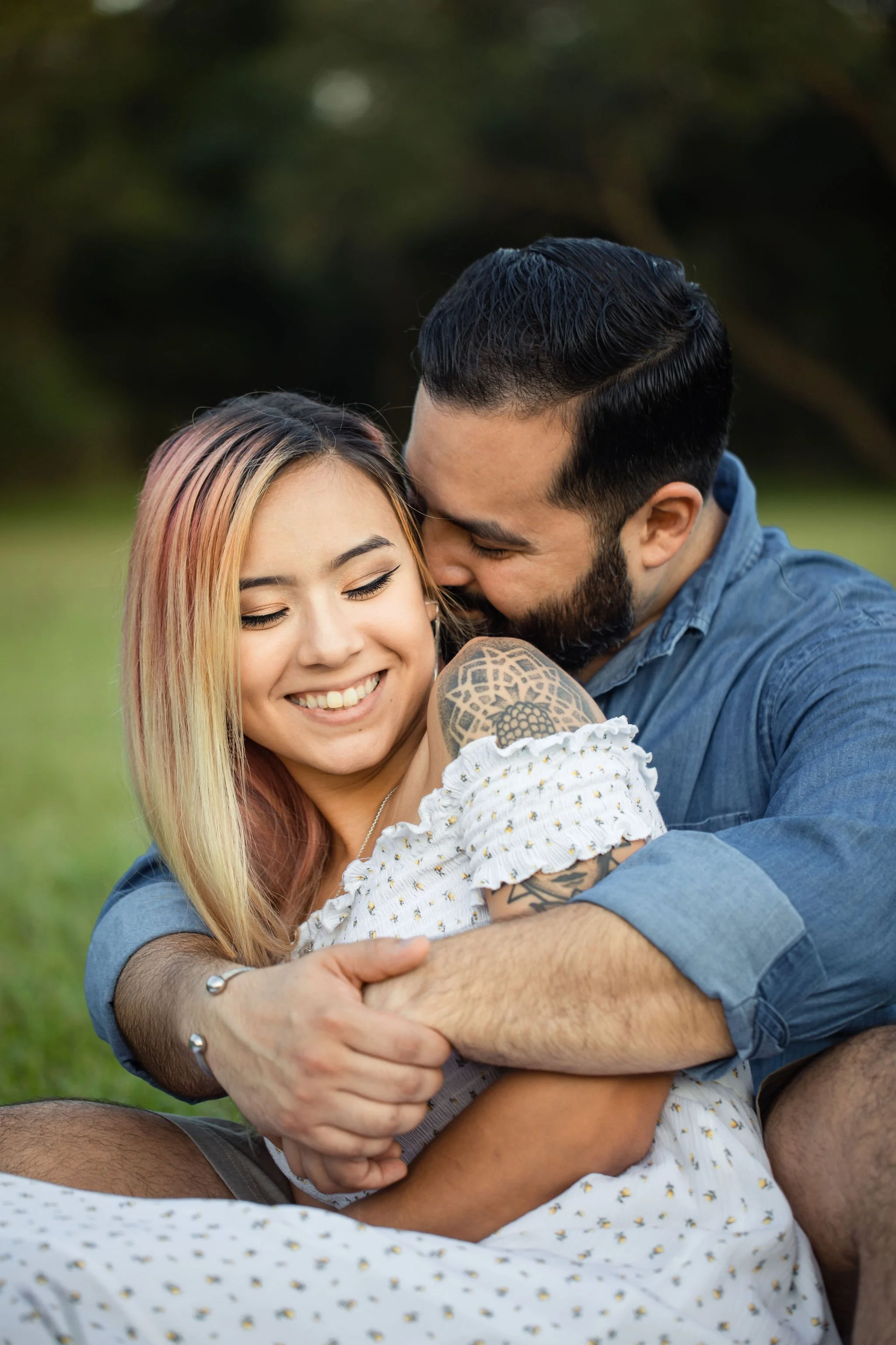Couple hugging and smiling outdoors with a woman with pink and blonde hair and a man with dark hair and a beard, in a grassy area.