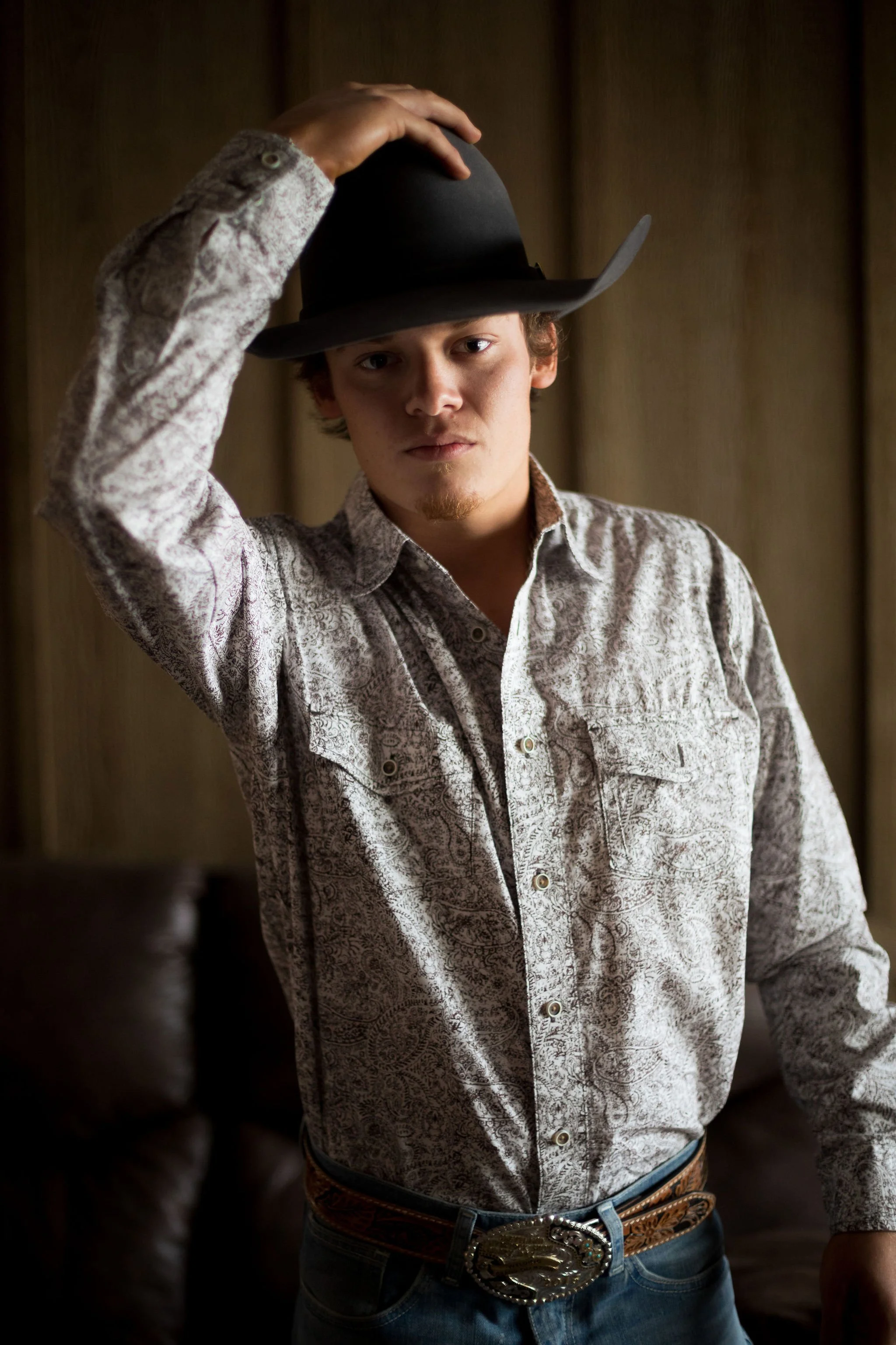 A young man wearing a cowboy hat, button-up shirt, and cowboy belt, standing indoors with wooden paneling in the background.
