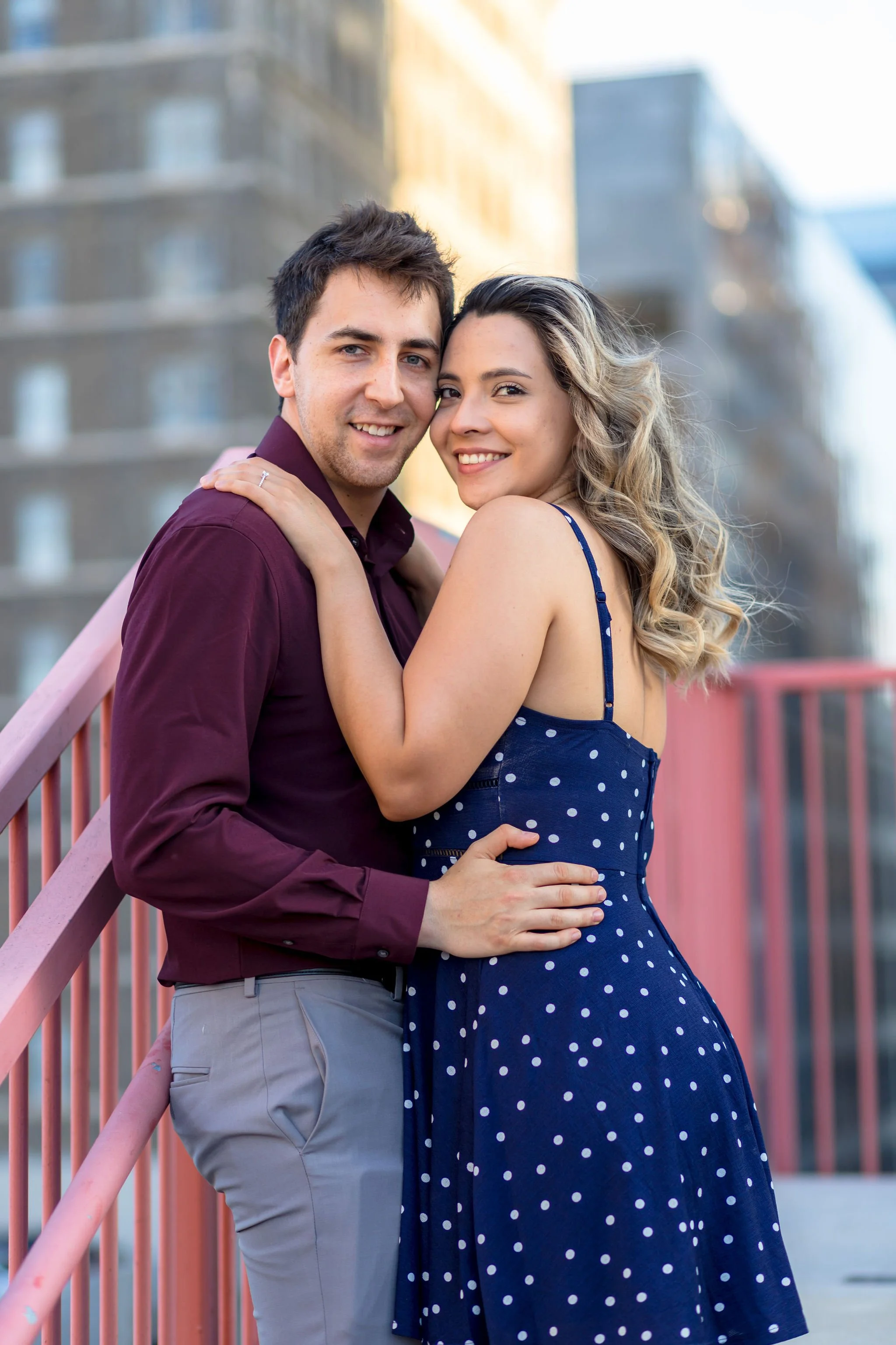 A young couple smiling and embracing on a pink bridge with city buildings in the background.