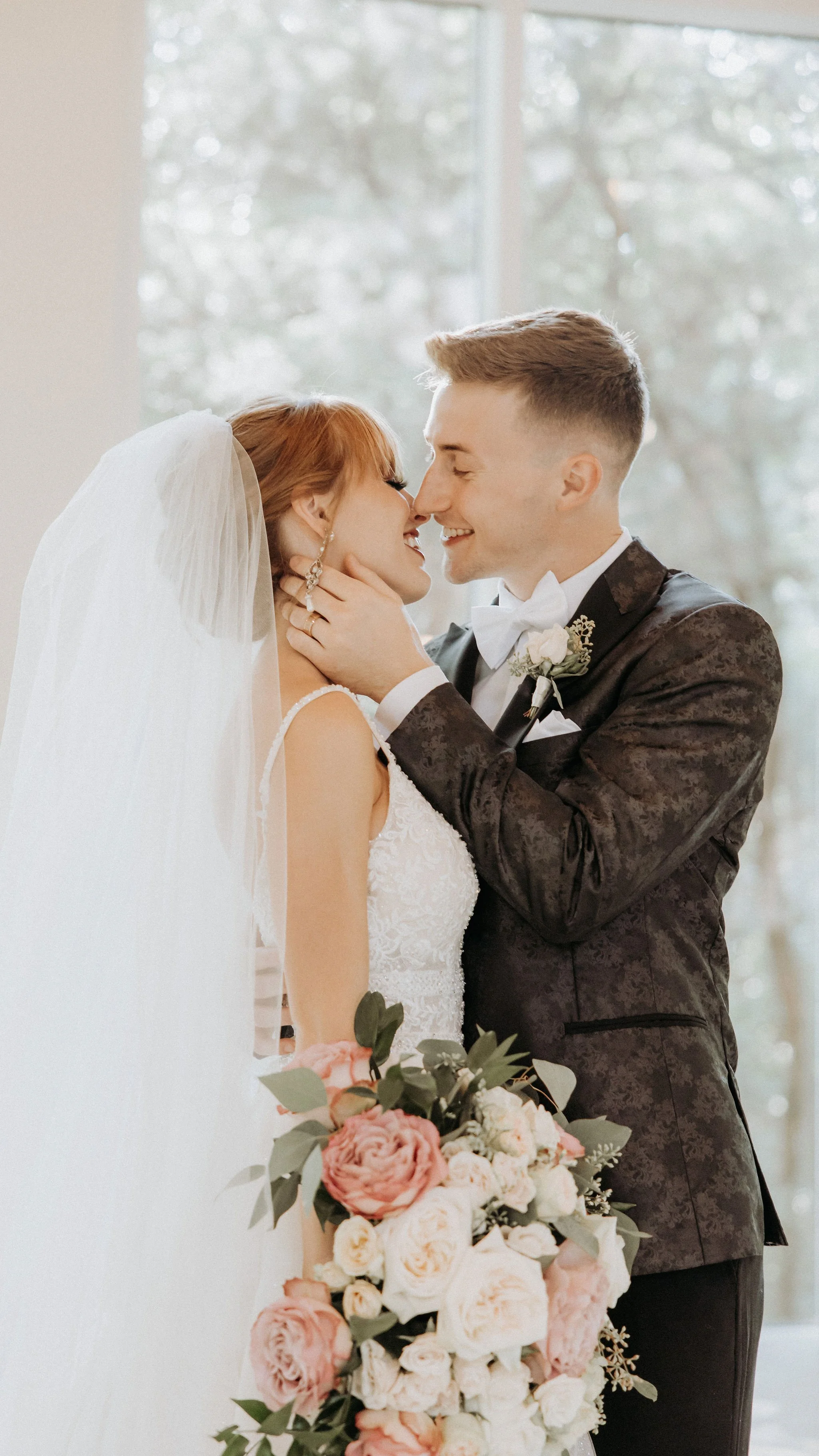 A bride and groom share an intimate moment, smiling with their foreheads touching, inside a bright room with large windows. The bride has red hair, wears a white wedding gown and veil, and holds a bouquet of pink and white roses. The groom wears a da