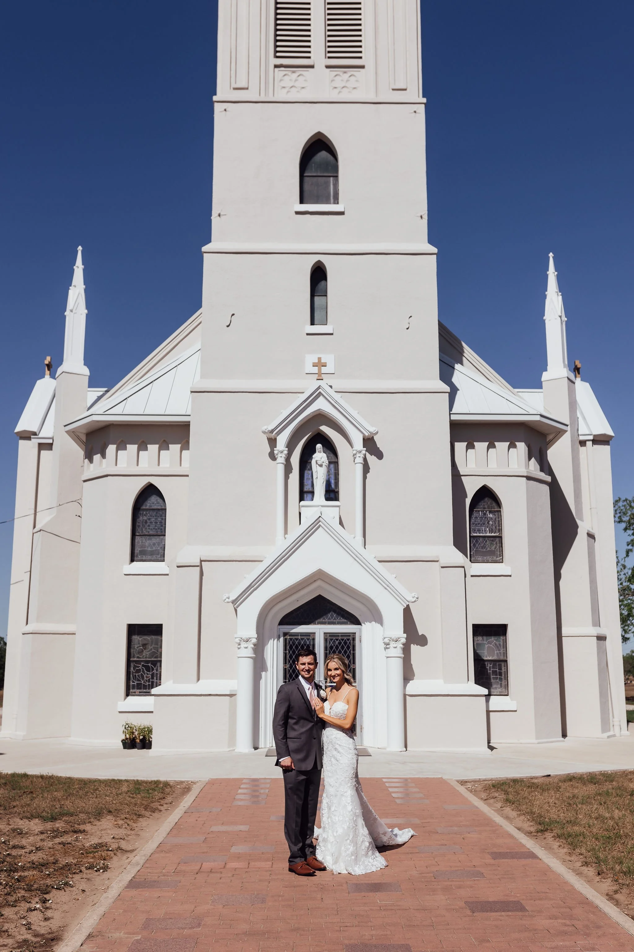 A newlywed couple in wedding attire standing on a brick pathway in front of a white church with gothic windows and spires under a clear blue sky.