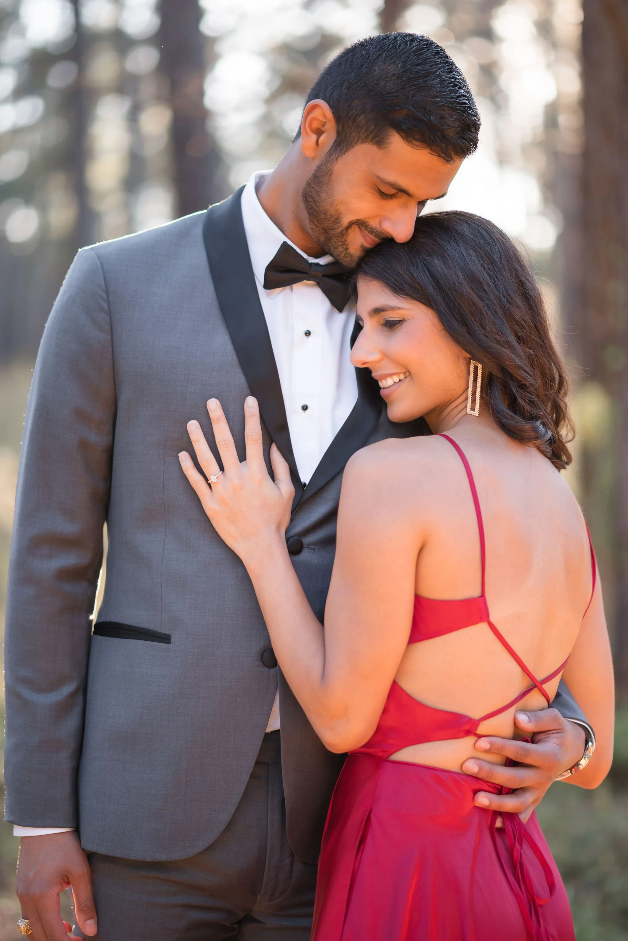 A romantic couple embracing outdoors, dressed elegantly with the man in a tuxedo and the woman in a red dress, smiling and touching each other affectionately.