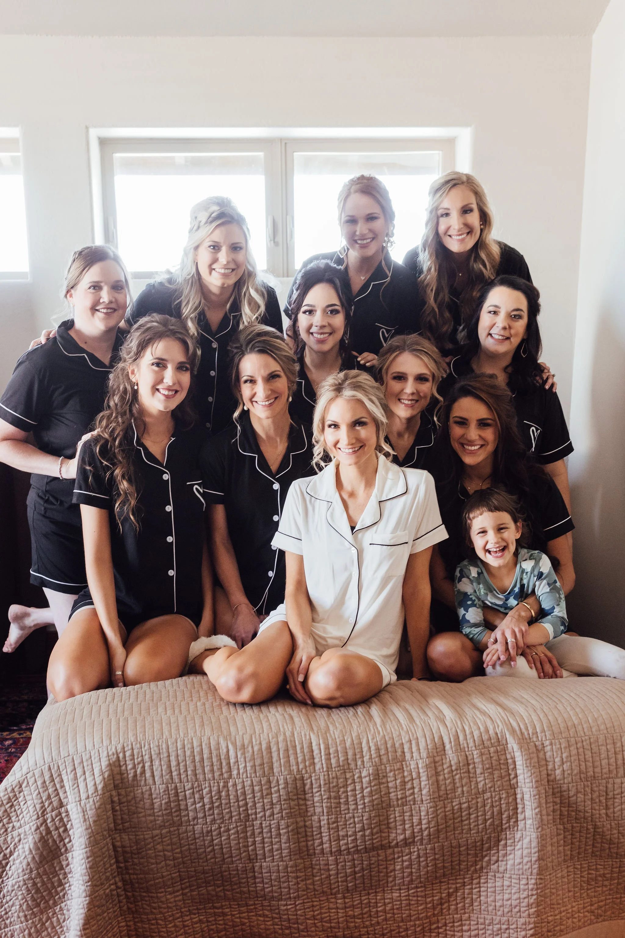 A group of women and one young girl in pajamas sitting and standing on a bed in a bright room.