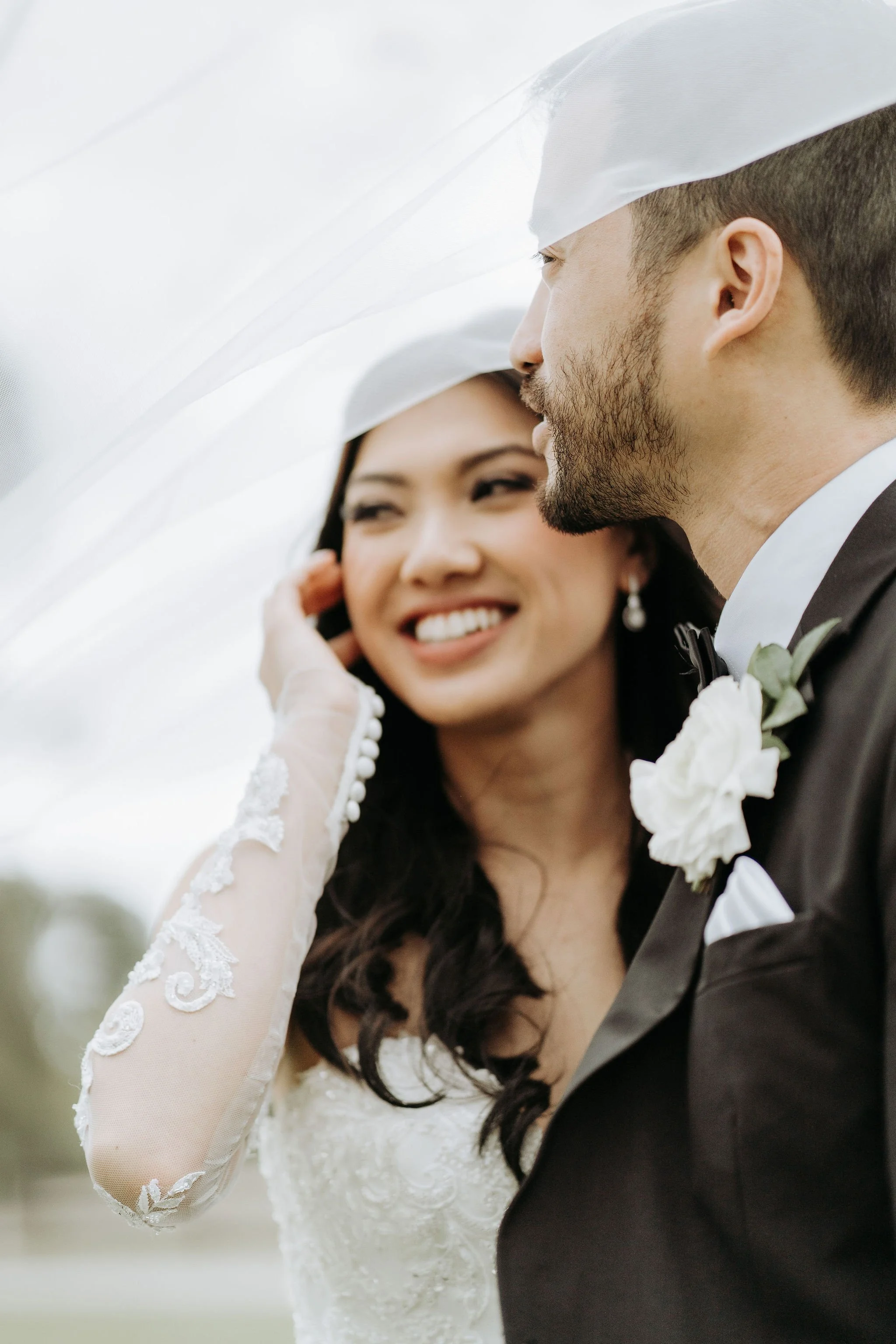 A bride and groom on their wedding day, standing outdoors under a white veil, smiling at each other.