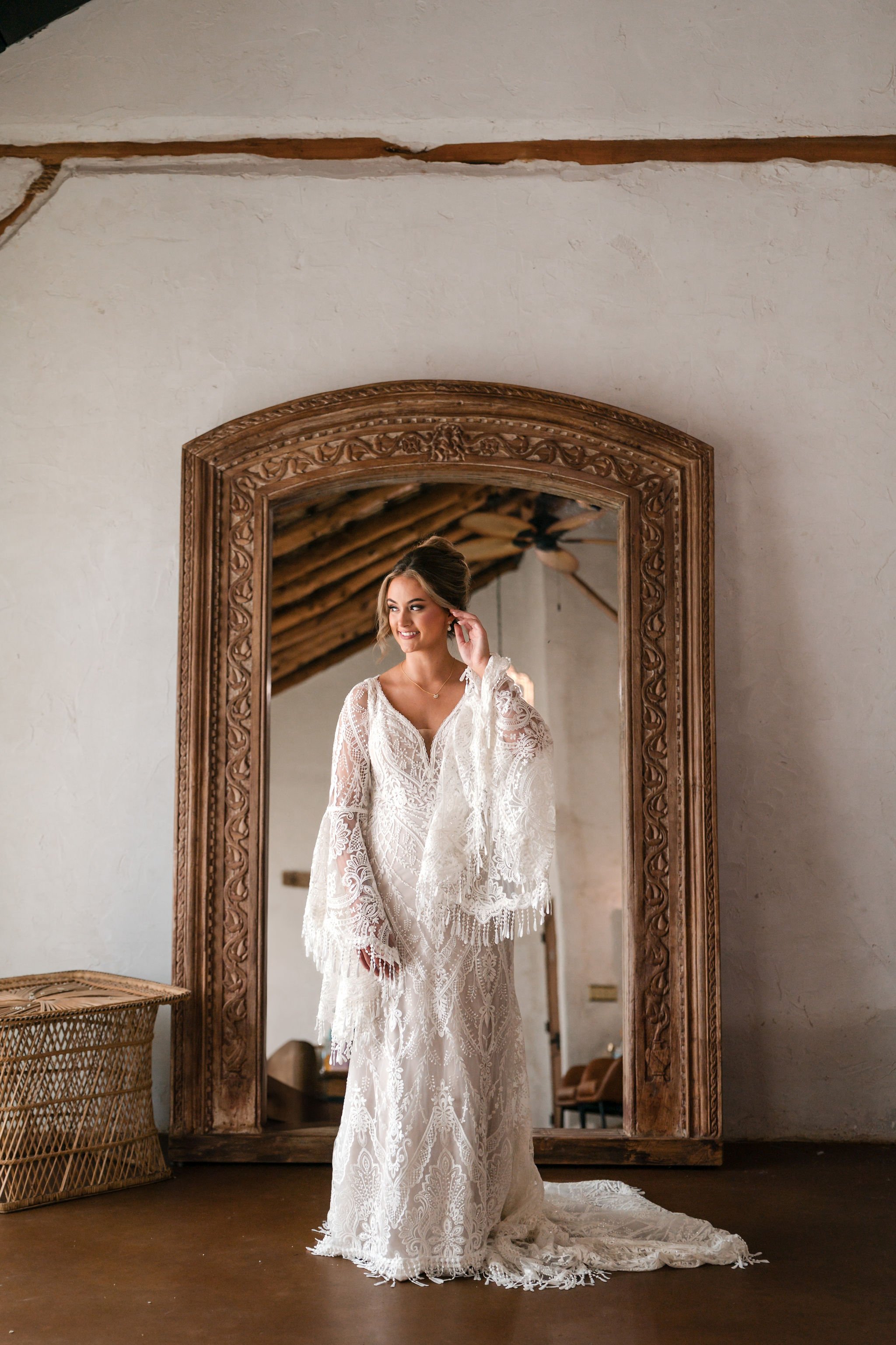 A bride in a white lace wedding dress stands in front of a large, ornate wooden framed mirror. She is smiling and adjusting her earring inside a rustic room with a sloped wooden ceiling and ceiling fan.