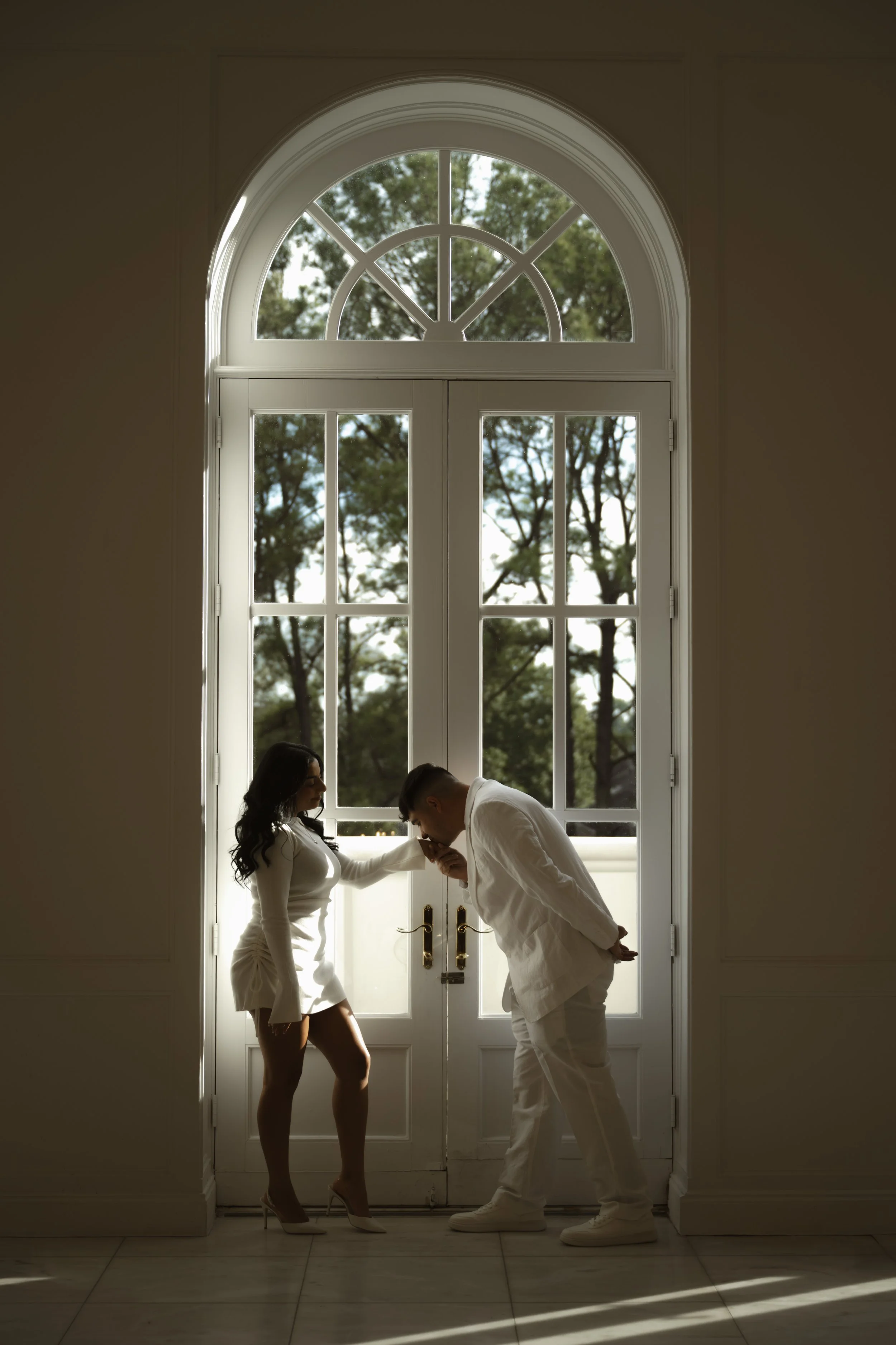 a couple is taking romantic engagement pictures in a white building and the man is kissing her hand 