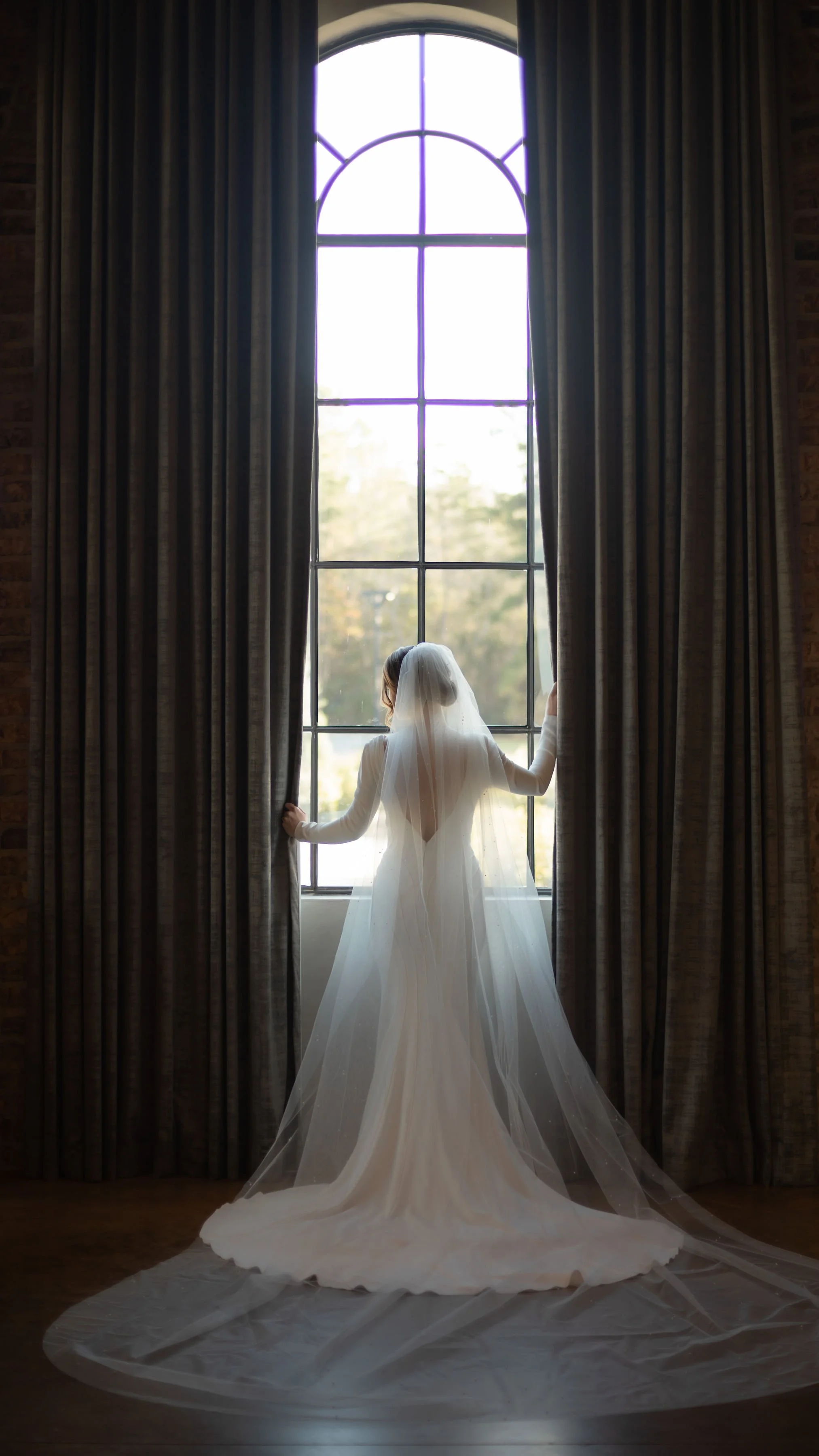 A bride in a white wedding dress and veil stands by a large window with dark curtains, looking outside.