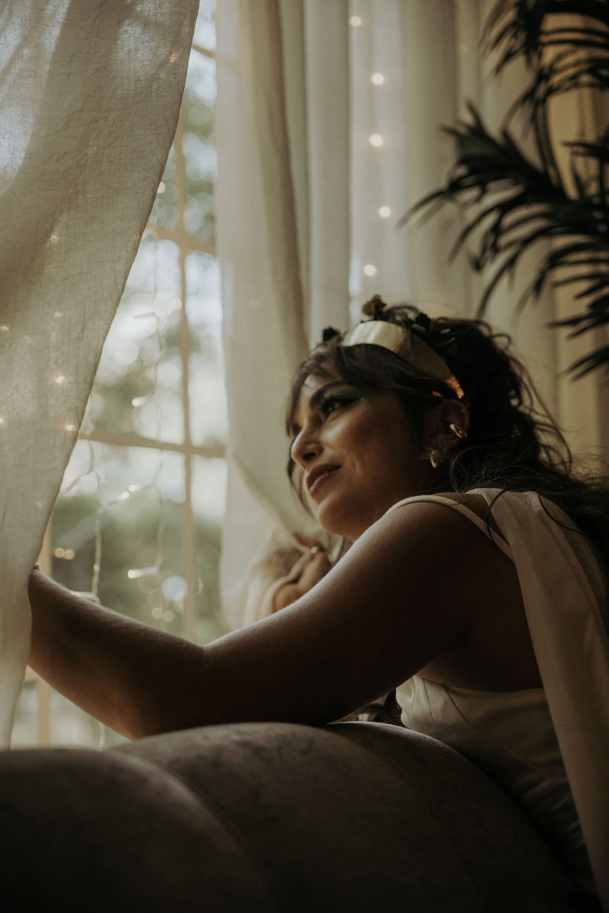 A woman with dark, wavy hair and a gold headband looks out a window, with sunlight and curtains in the background.