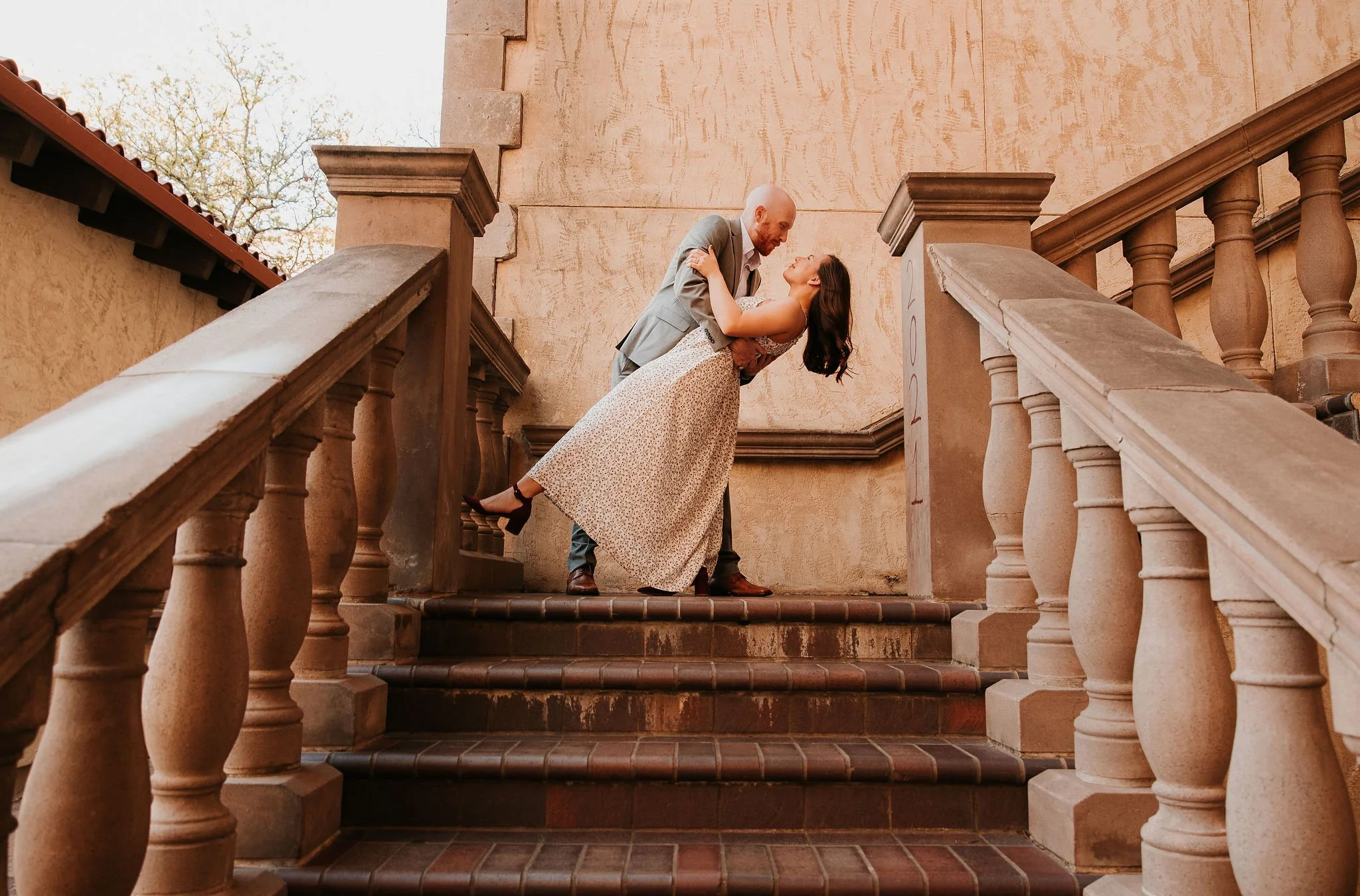 A couple dressed in formal attire dancing on an outdoor staircase with beige stone walls and wooden balustrades.