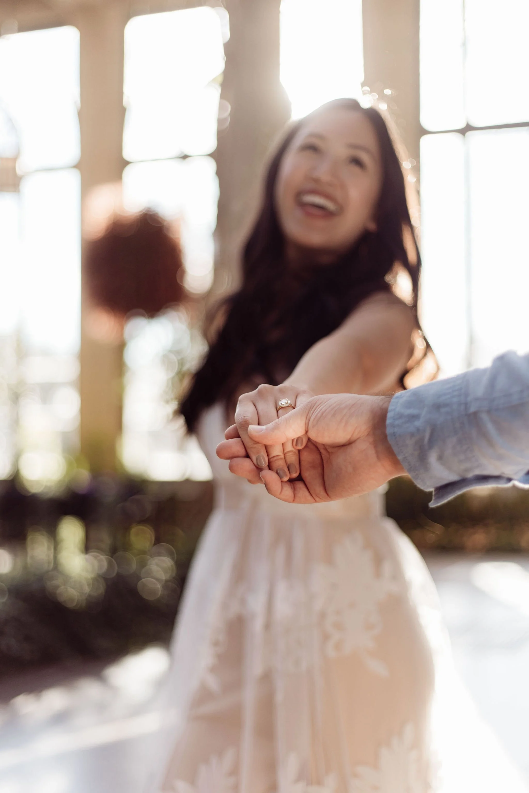A woman wearing a wedding dress is holding hands with a person in a long-sleeve shirt, smiling, with sunlight streaming through the windows in the background.