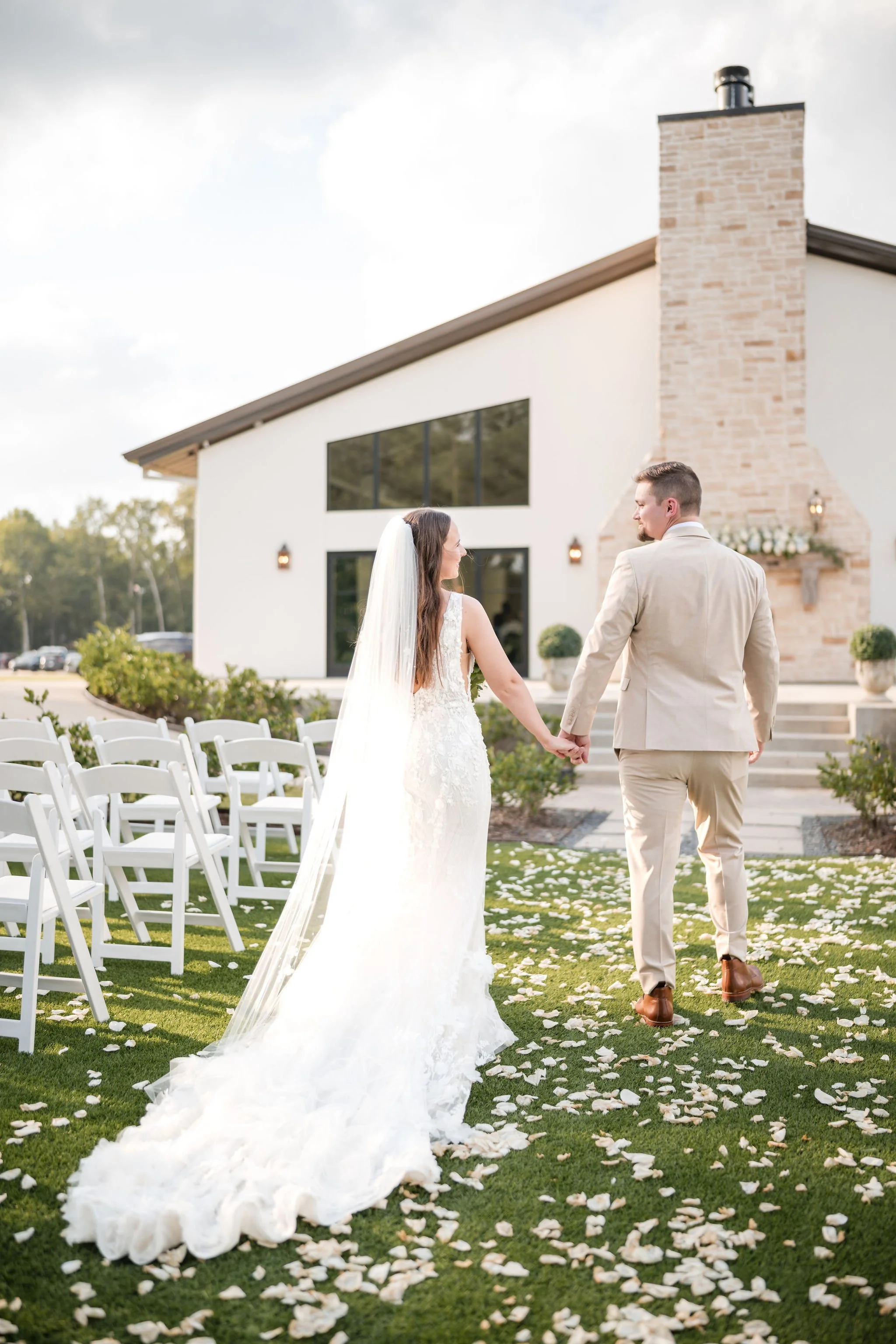 A bride and groom holding hands and walking away from the camera on an outdoor wedding aisle with flower petals scattered on the grass, a modern white building with large windows and a brick chimney in the background.