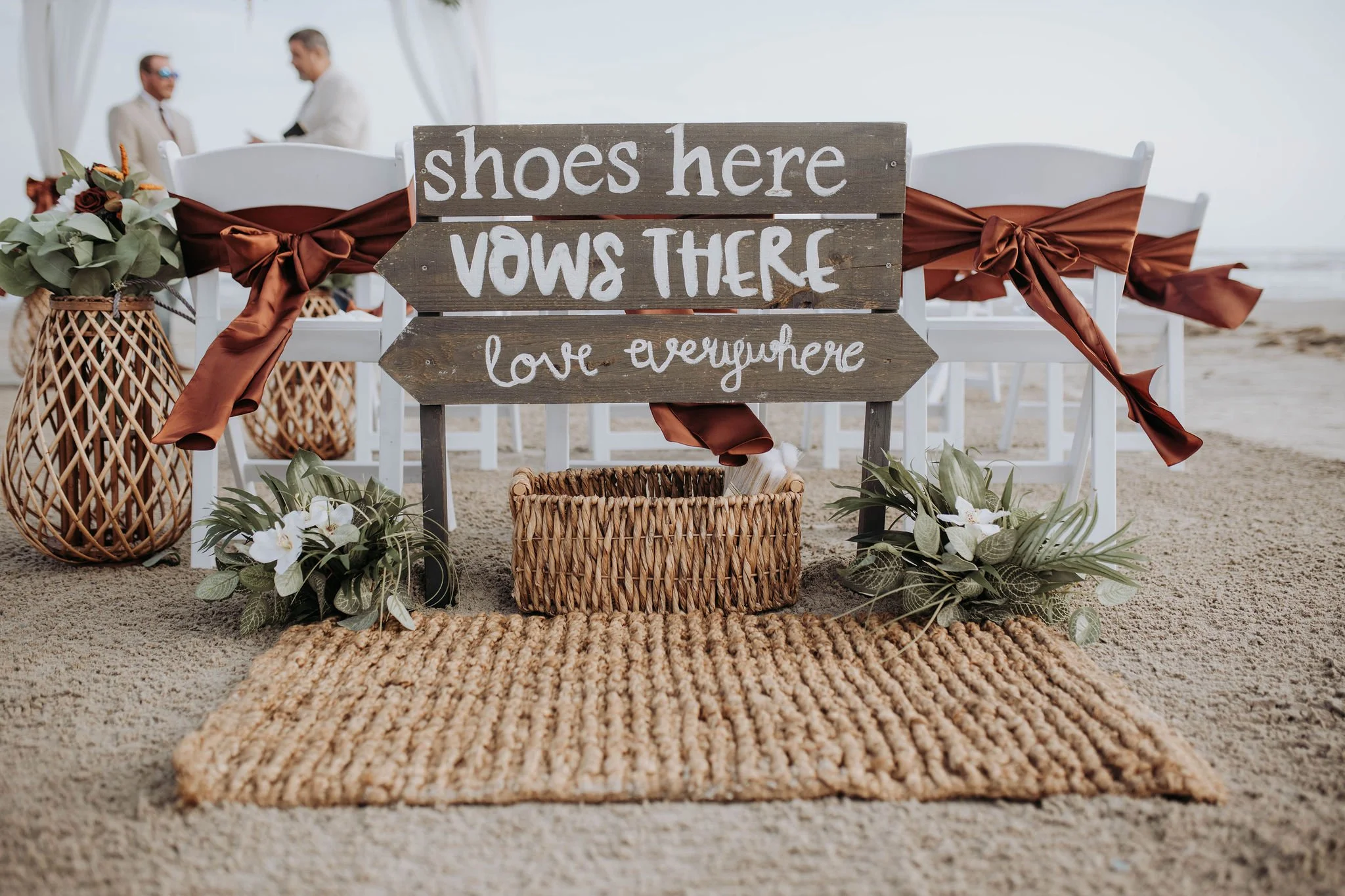 Beach wedding setup with a wooden sign that says "shoes here, vows there, love everywhere" flanked by flowers and decorations, with two people in the background near the ocean.
