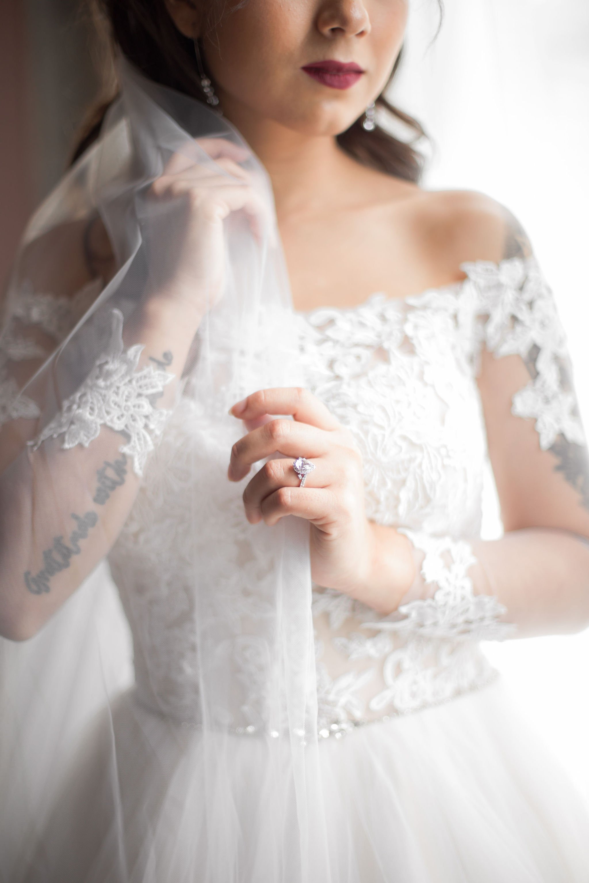 A bride in a white lace wedding dress holding a wedding veil and displaying an engagement ring.