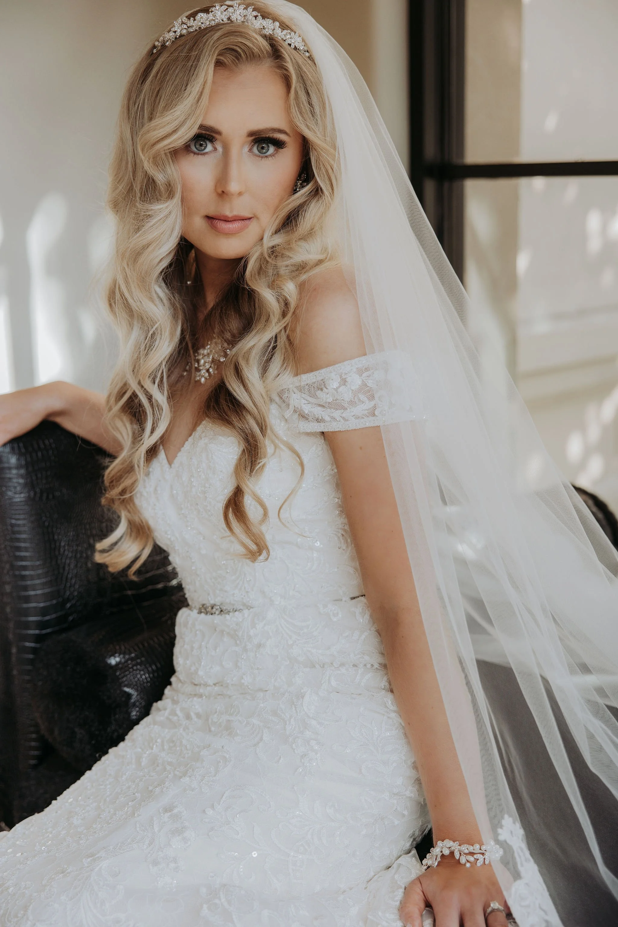 A bride with long, wavy blonde hair wearing a white wedding gown, veil, and jewelry, sitting indoors near a window.