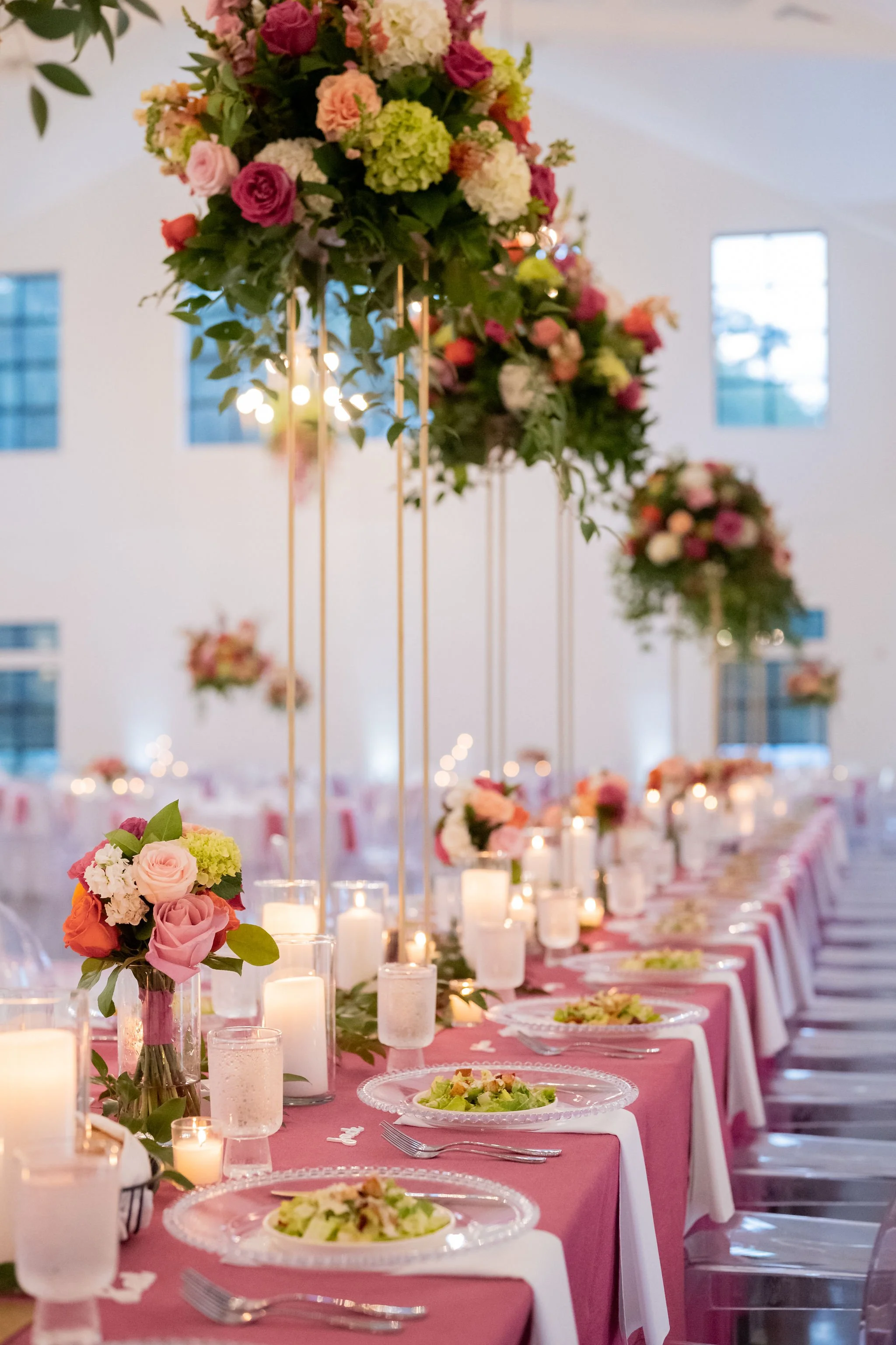 Elegant banquet table setting with pink tablecloths, floral centerpieces in pink, white, and green, and lit candles. The table is set with glass plates, silverware, and salads, in a decorated event hall with high ceilings and large windows.