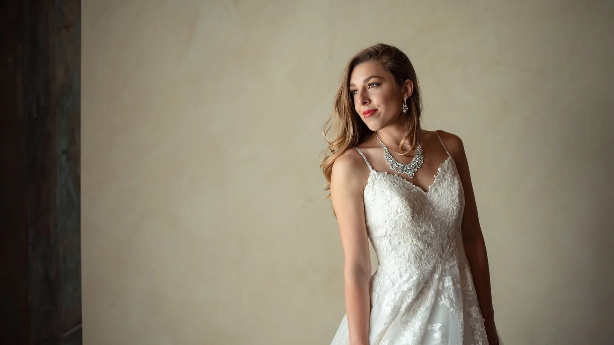 A young woman in a wedding dress with lace details, wearing earrings and a necklace, gazing to her left, standing against a neutral background.