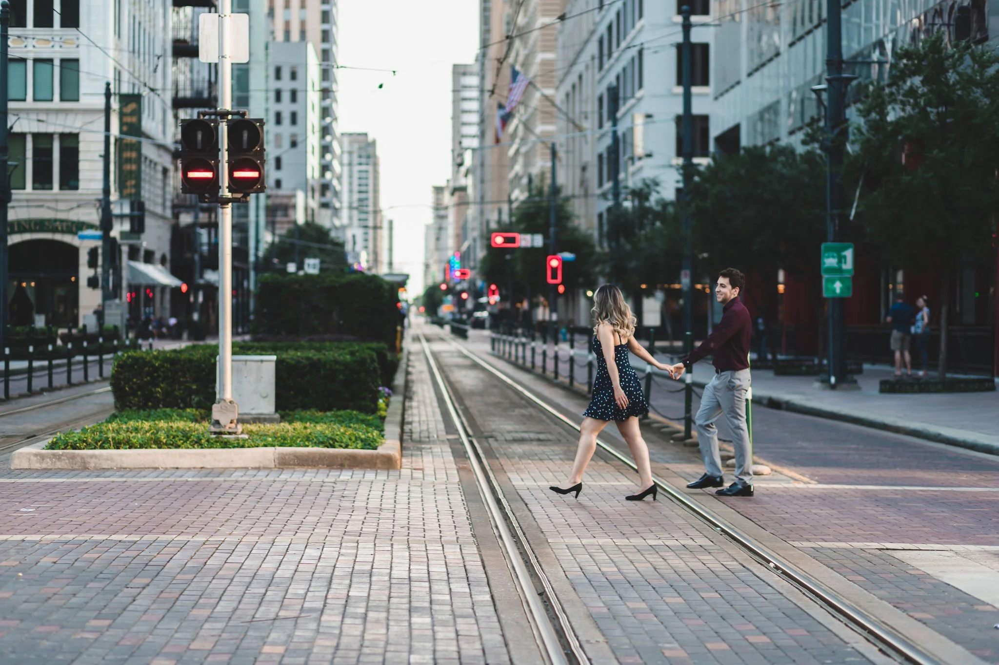 A couple holding hands and crossing the street on tram tracks in an urban area with tall buildings and traffic signals.