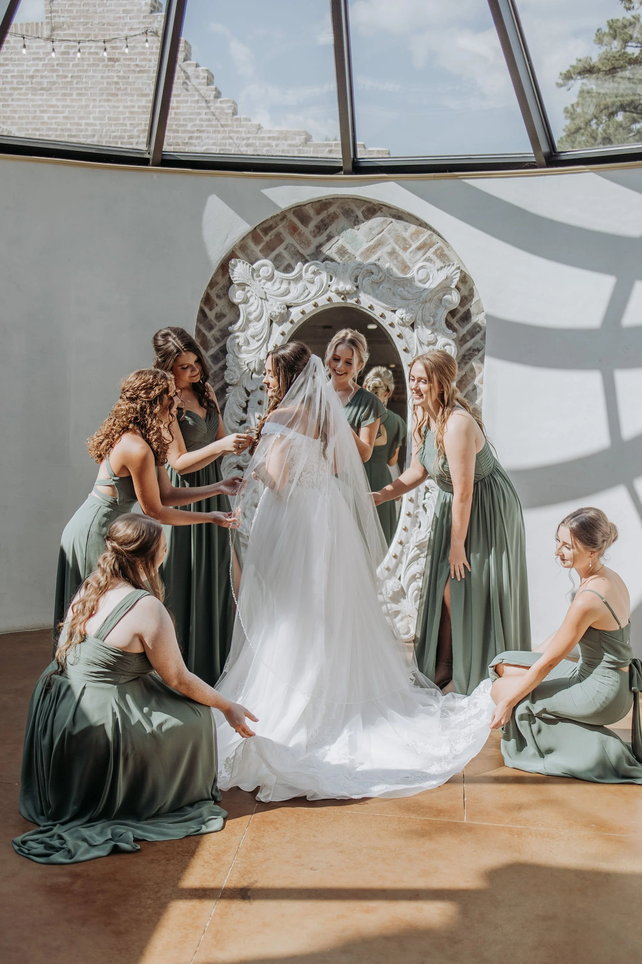 Bride in white wedding gown with veil surrounded by her bridesmaids in green dresses, helping her in a room with ornate mirror and large windows casting shadows.