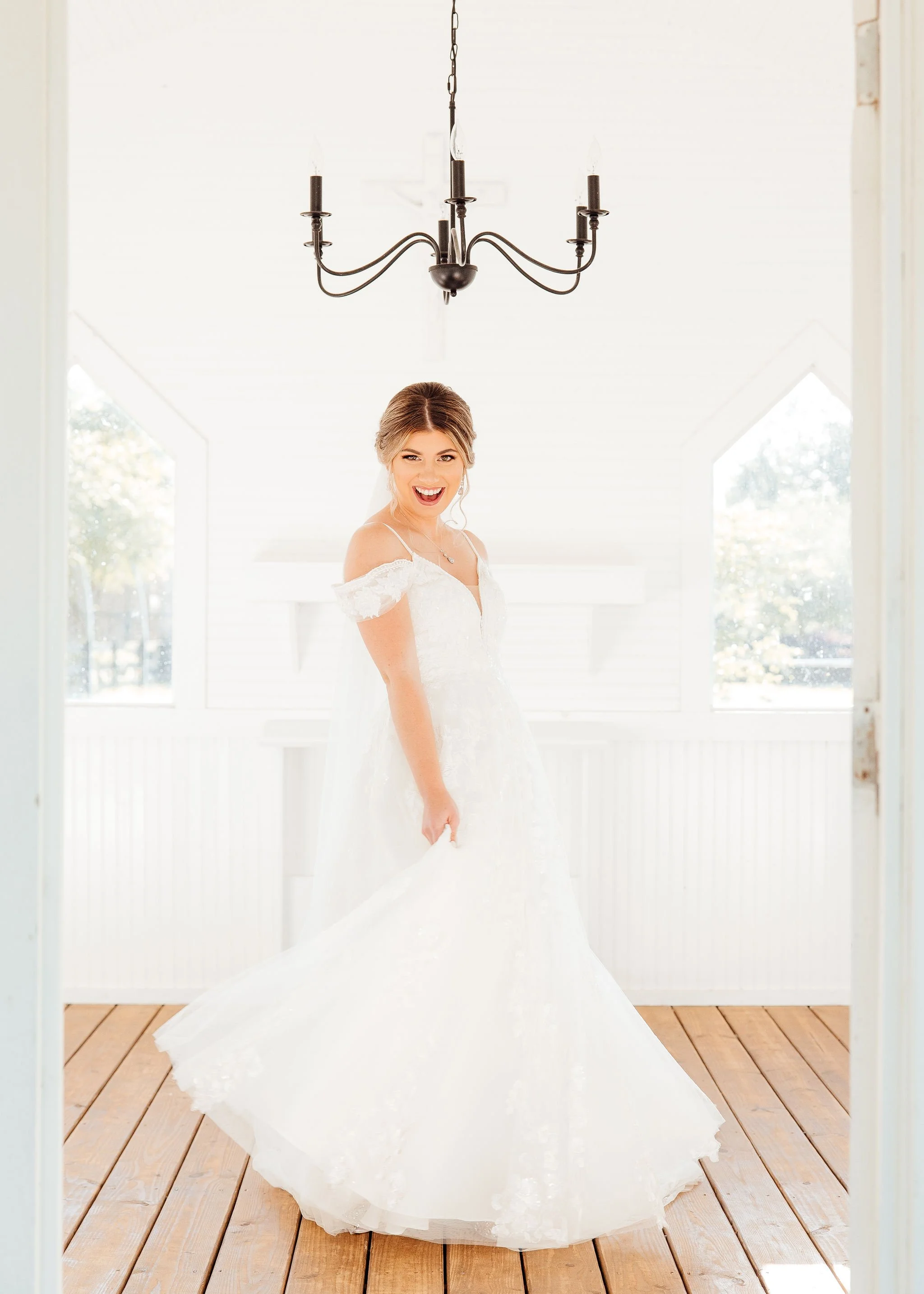 A bride in a white wedding dress smiling inside a bright room with wooden floors and large windows, hanging black chandelier overhead.