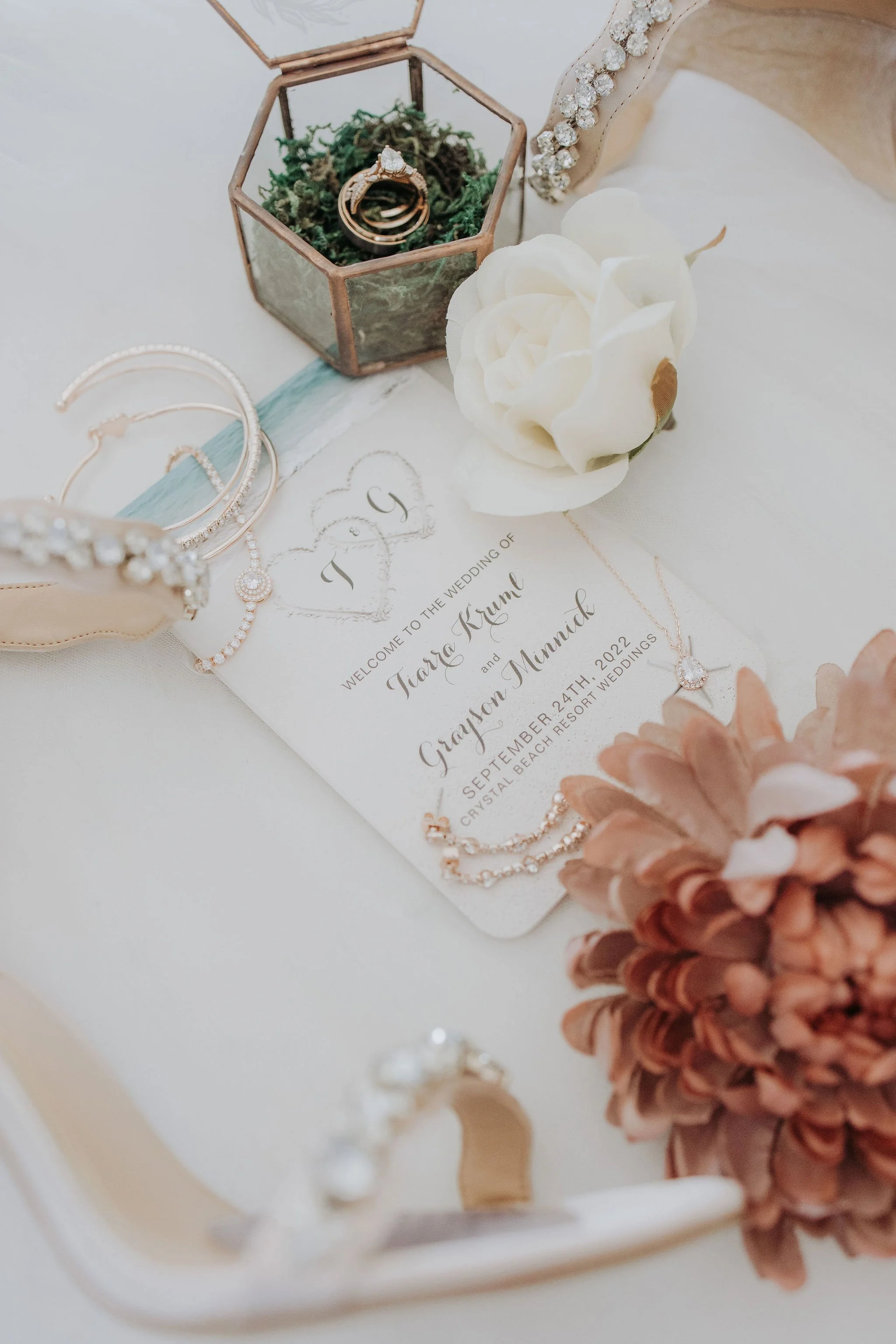Wedding invitation surrounded by jewelry, flowers, and a glass container on a white surface.