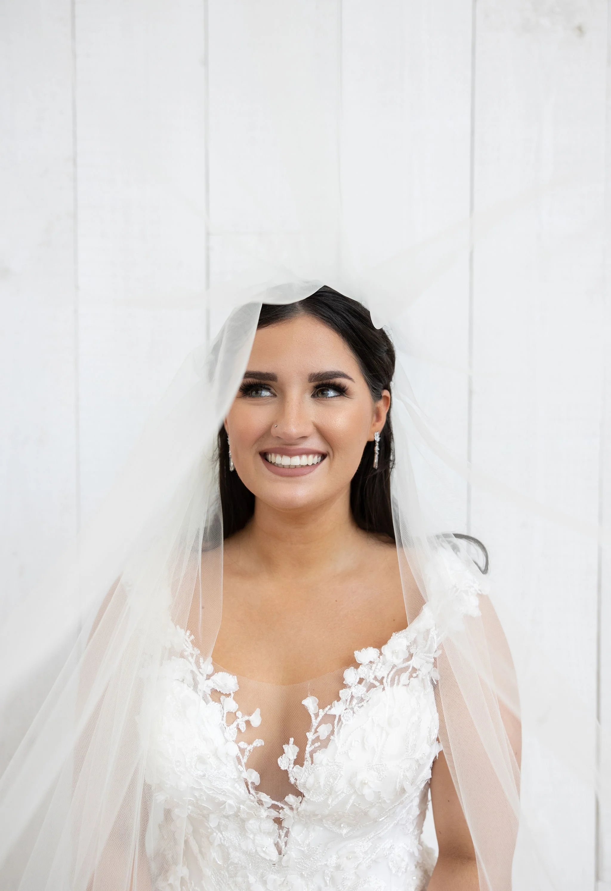 Bride in a white wedding dress and veil smiling and looking to the side, standing against a white wall.