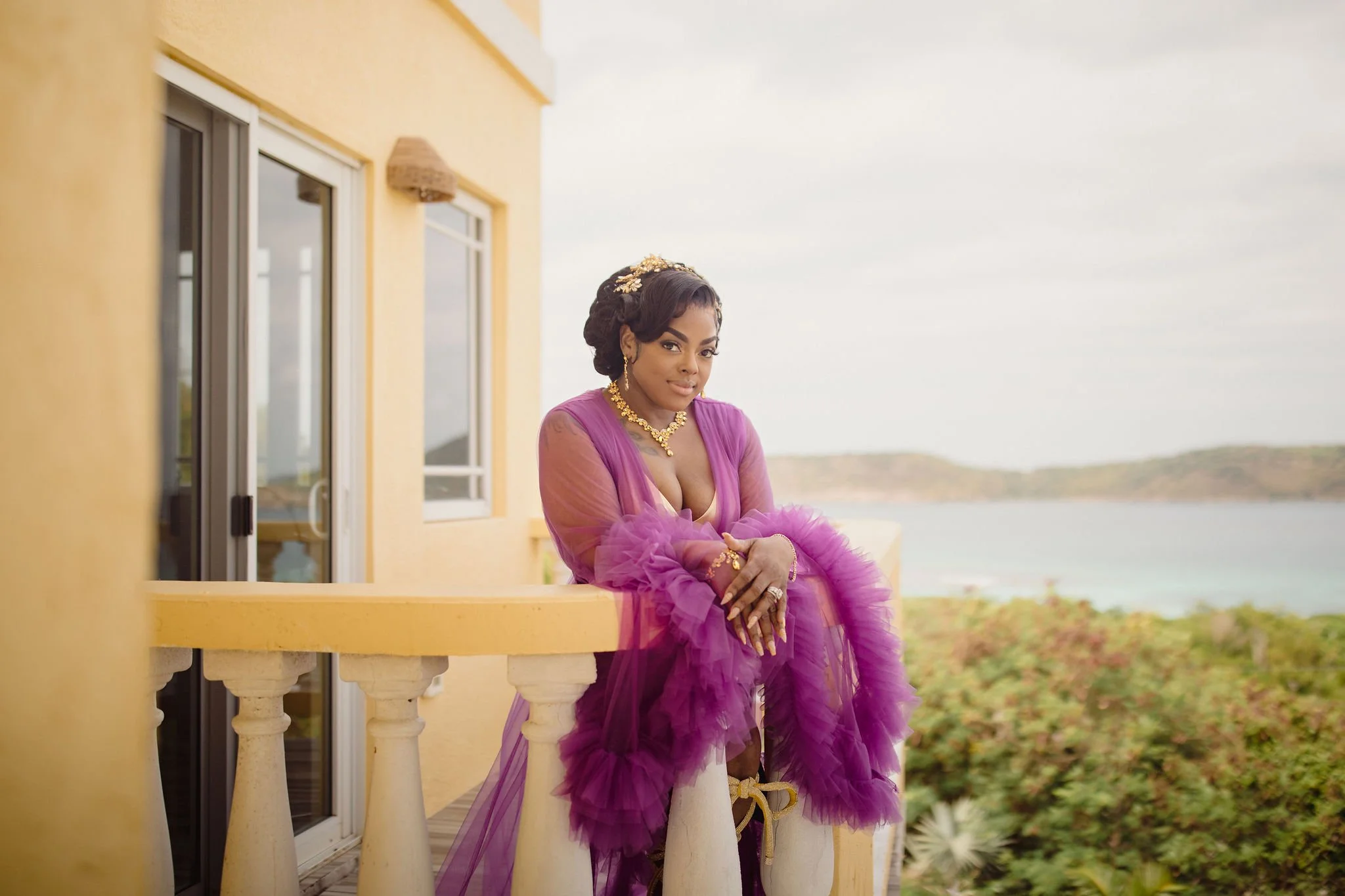 A woman in a purple tulle dress with gold jewelry, standing on a balcony overlooking water and hills.