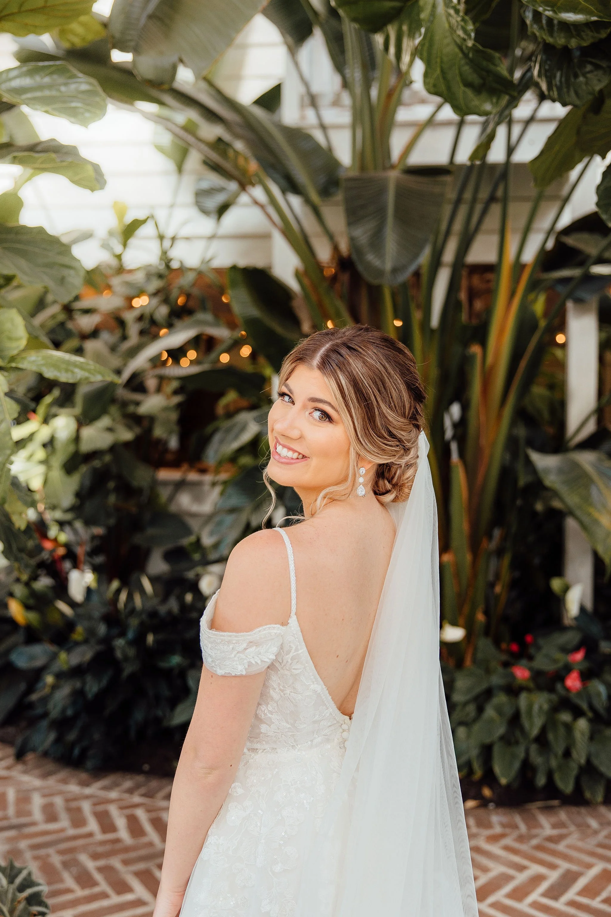 A bride in a wedding dress with a veil, smiling and looking over her shoulder, surrounded by lush green plants in a bright greenhouse or conservatory.