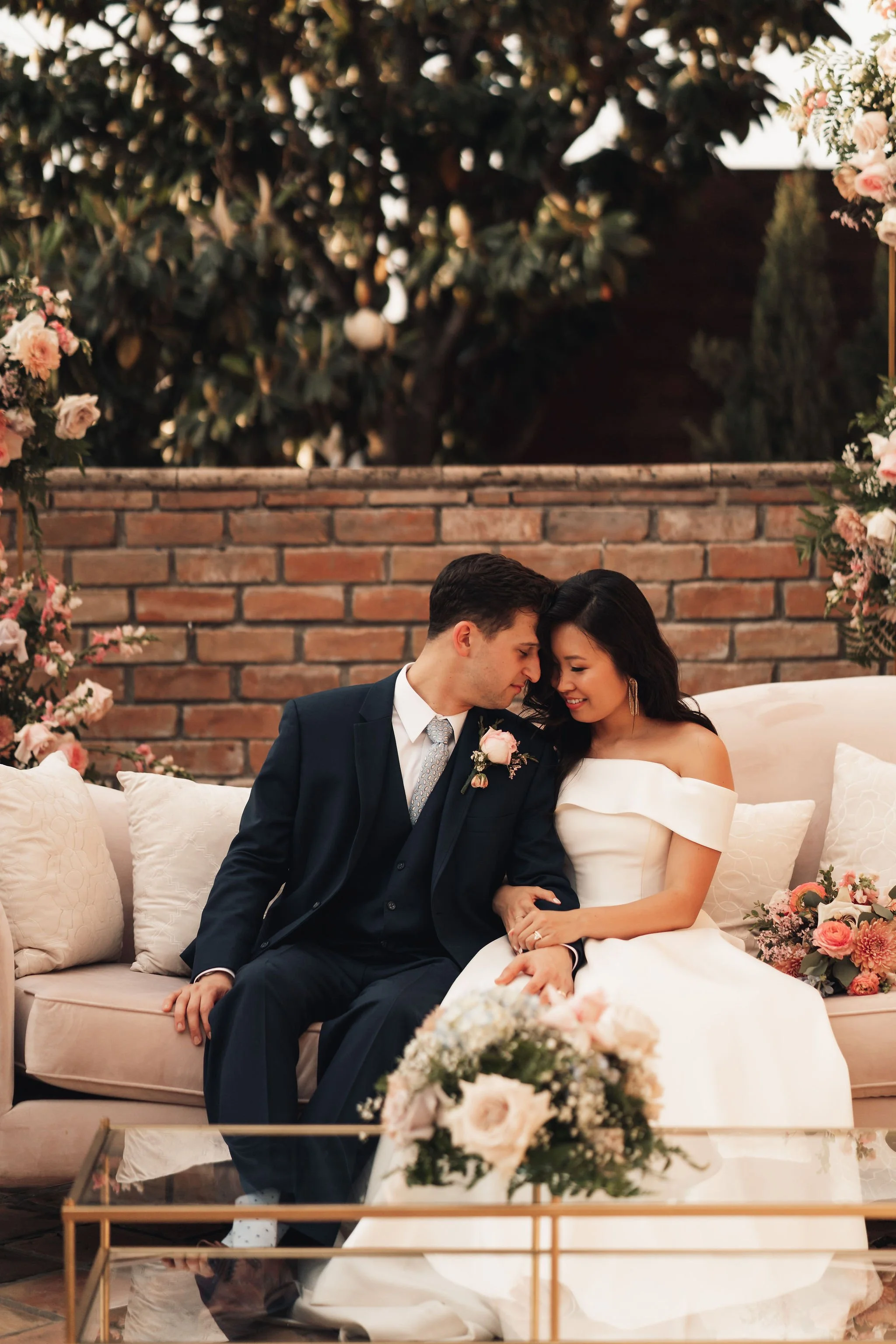A newlywed couple sitting close together on a cream-colored sofa, with their foreheads touching and smiling. The groom is dressed in a dark suit with a boutonniere, and the bride is wearing a white off-shoulder wedding gown. There are floral arrangem