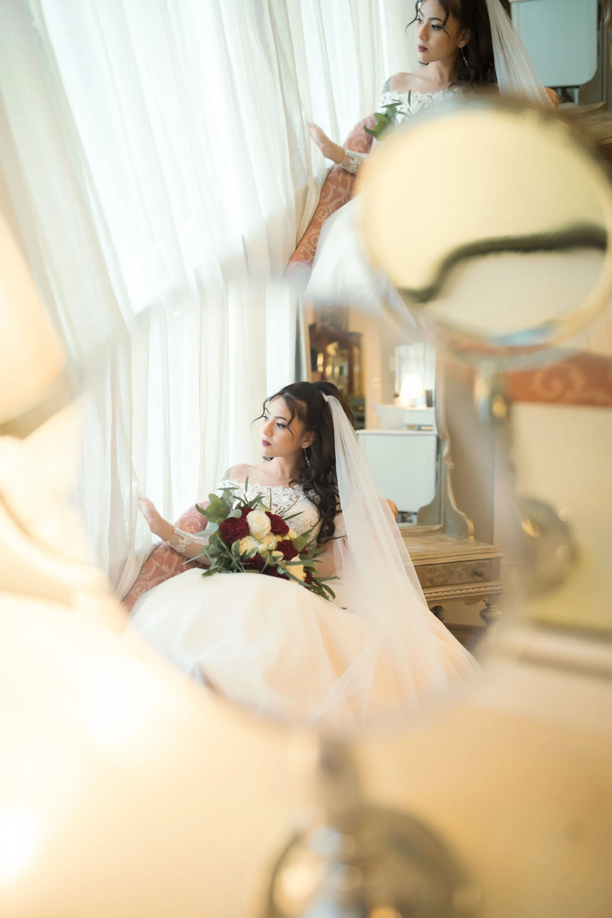 Bride sitting by a window with white curtains, holding a bouquet of red and white roses, reflected in a mirror.
