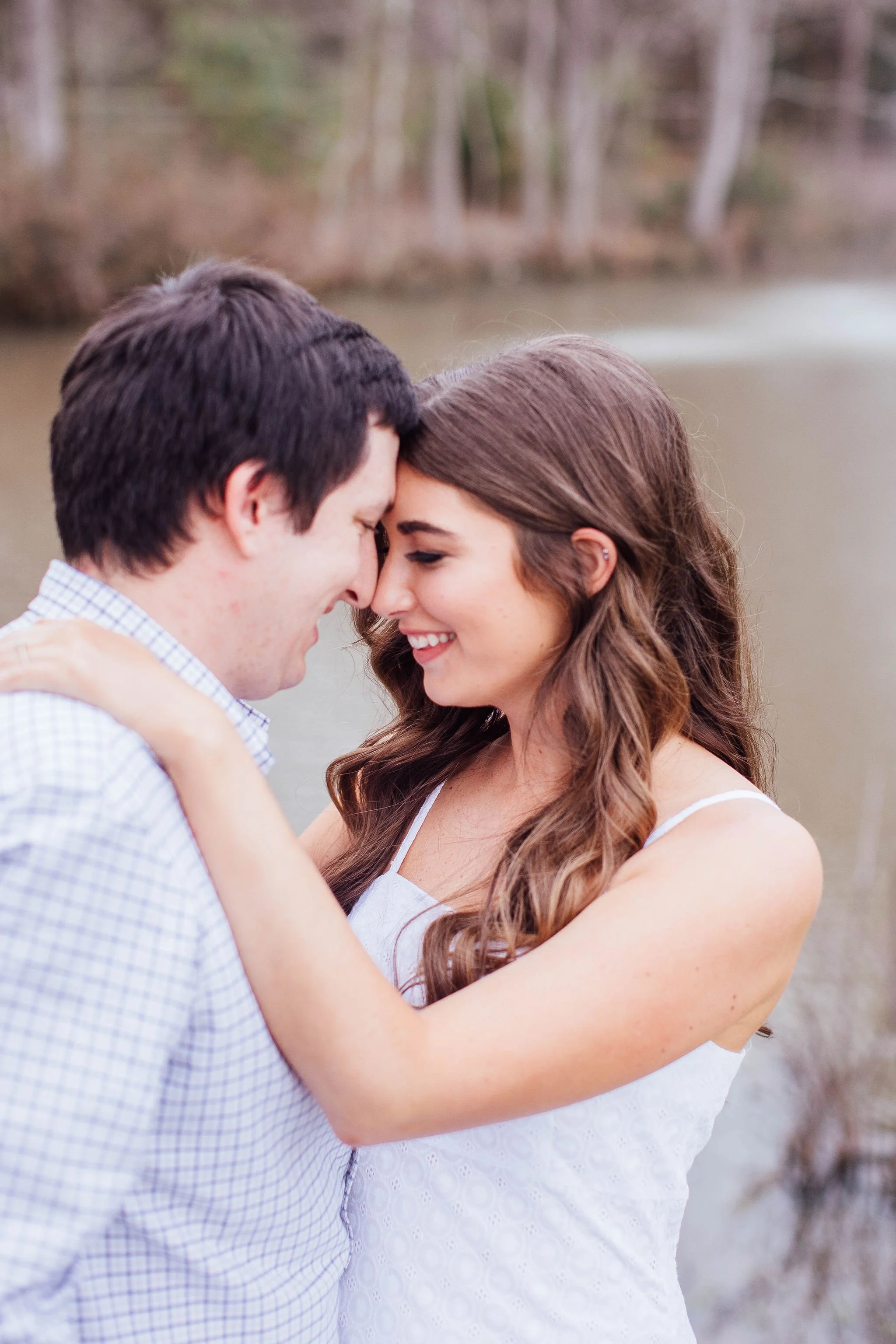 A couple embracing and touching foreheads near a body of water with trees in the background.