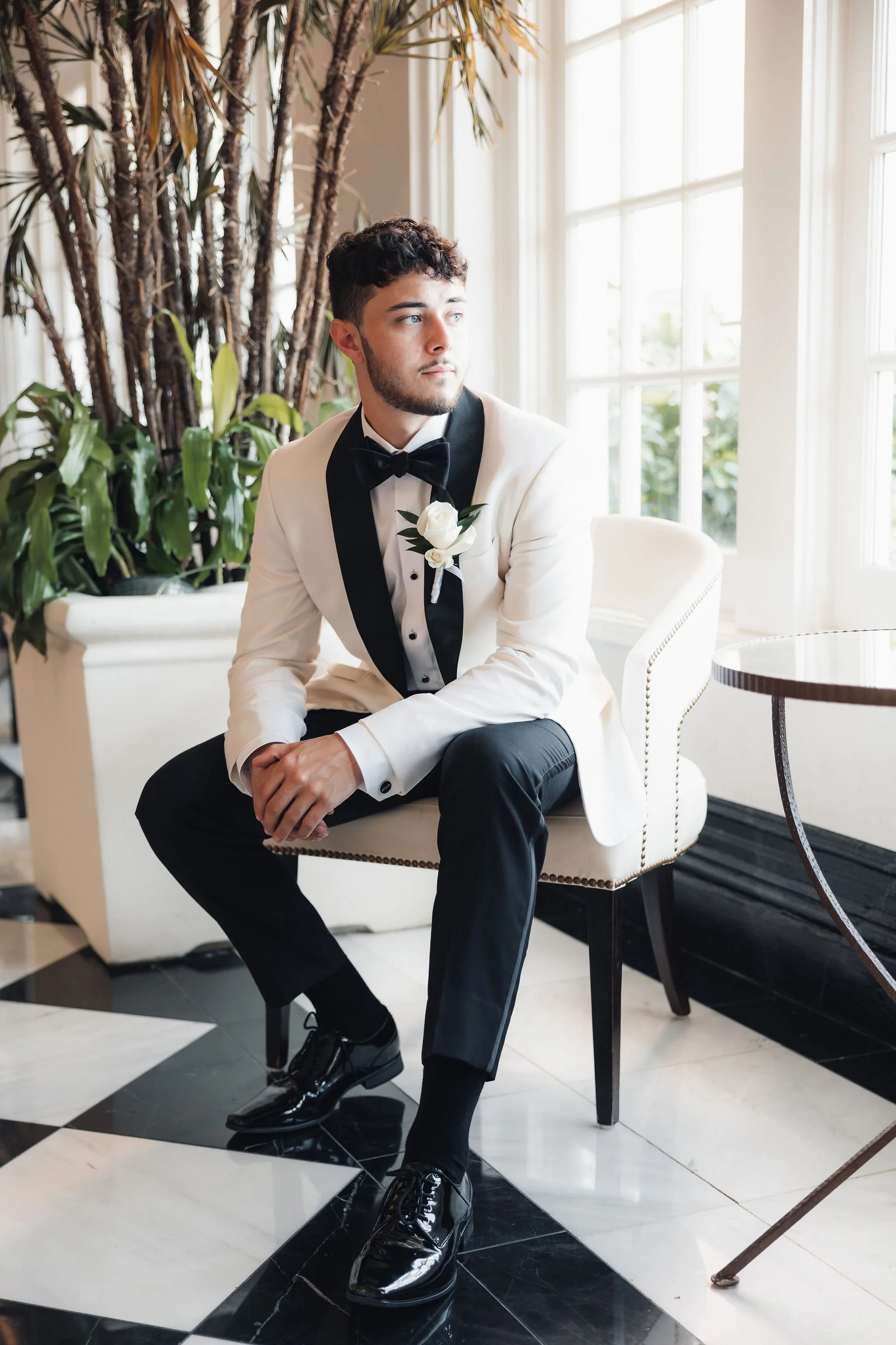 Man in a white tuxedo with black trim, black bow tie, and boutonniere, sitting on a cream-colored chair near large windows with sunlight, surrounded by plants and black-and-white tiled floor.