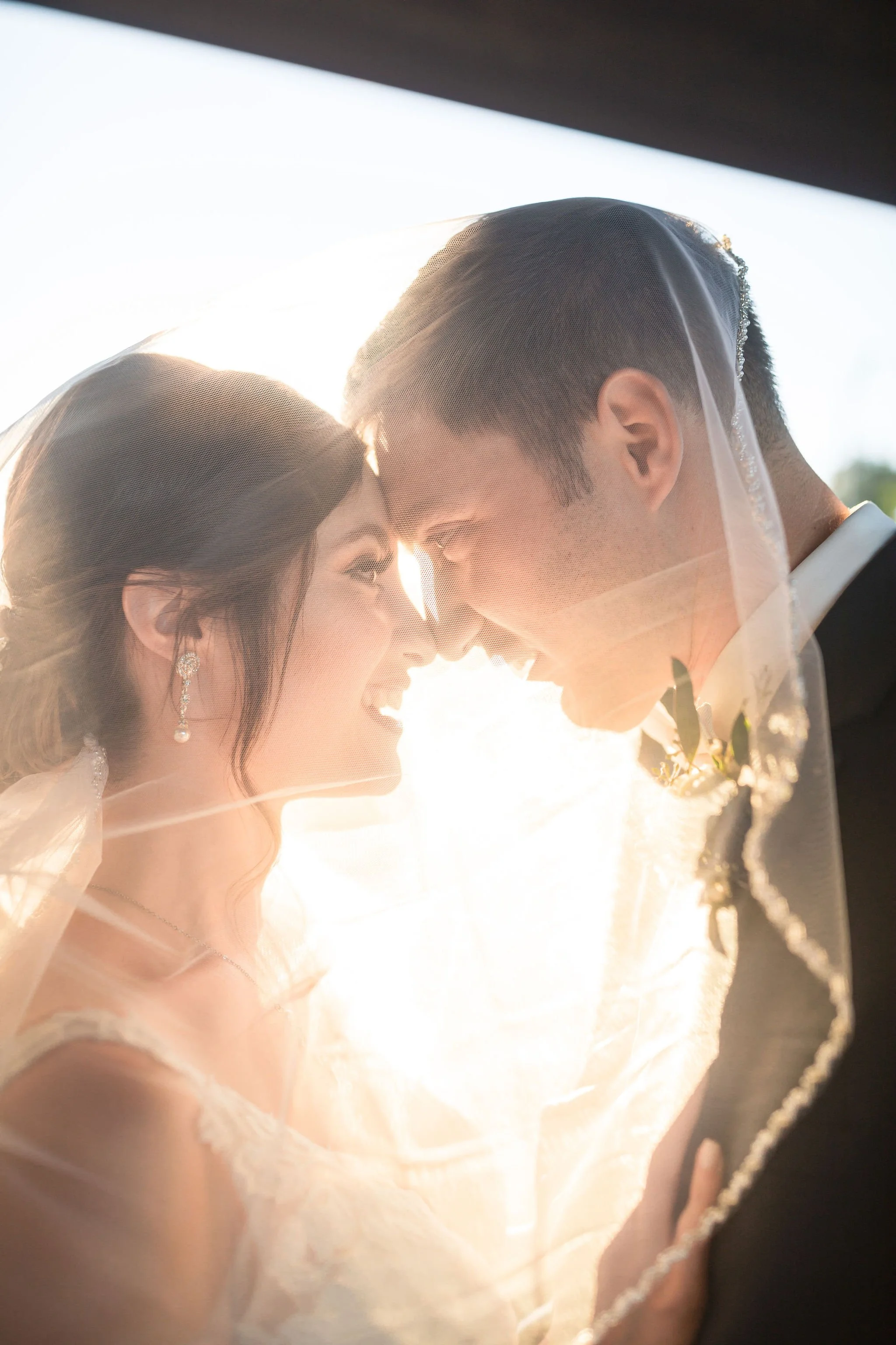 A bride and groom with their foreheads touching, smiling, inside a vehicle with sunlight shining through, during their wedding celebration.