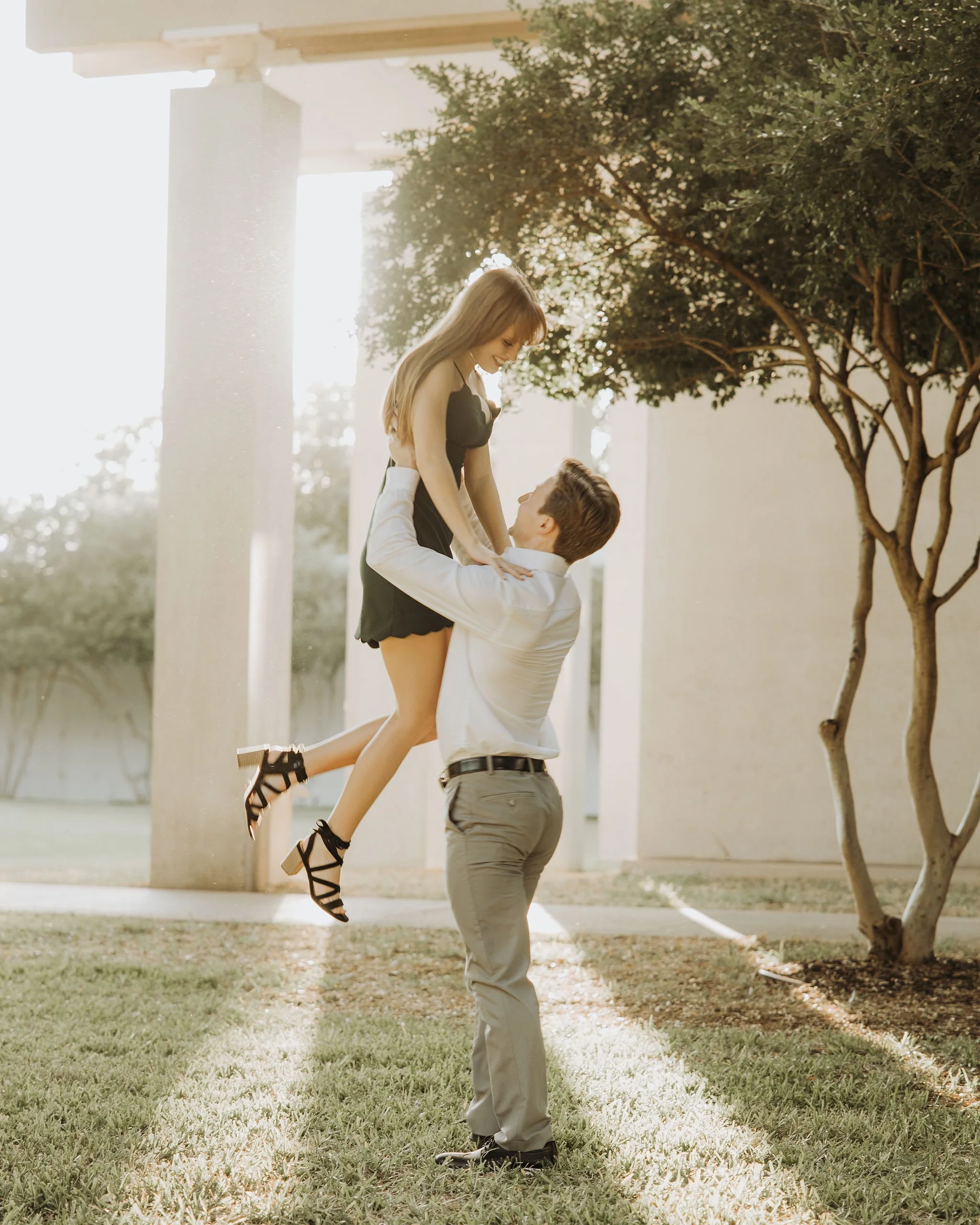 A man lifting a woman into the air outdoors near a tree, with sunlight creating a bright, warm atmosphere.
