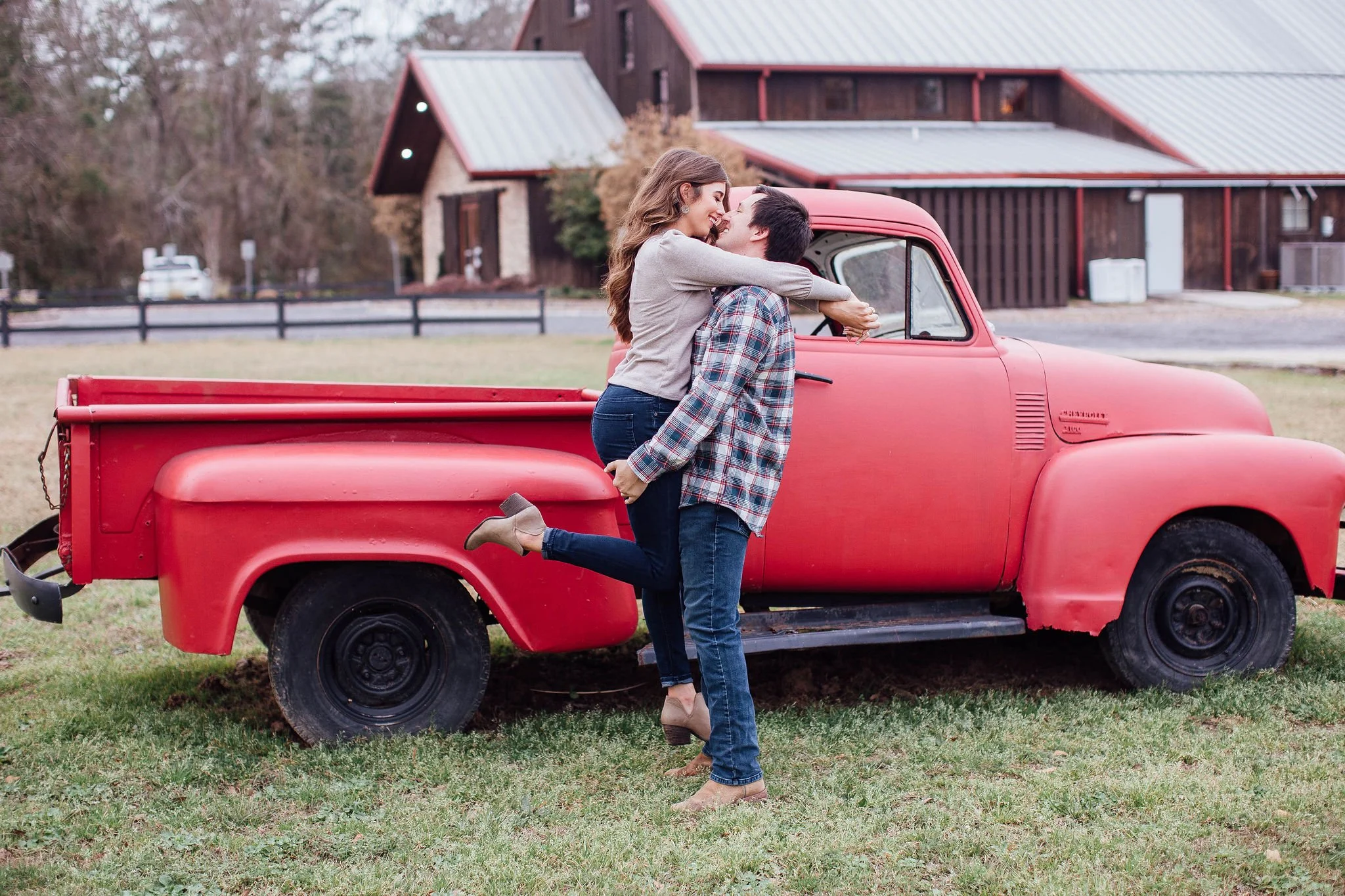 A couple is standing next to a vintage red pickup truck in a grassy area, with the woman sitting on the man's lap, smiling and embracing each other.