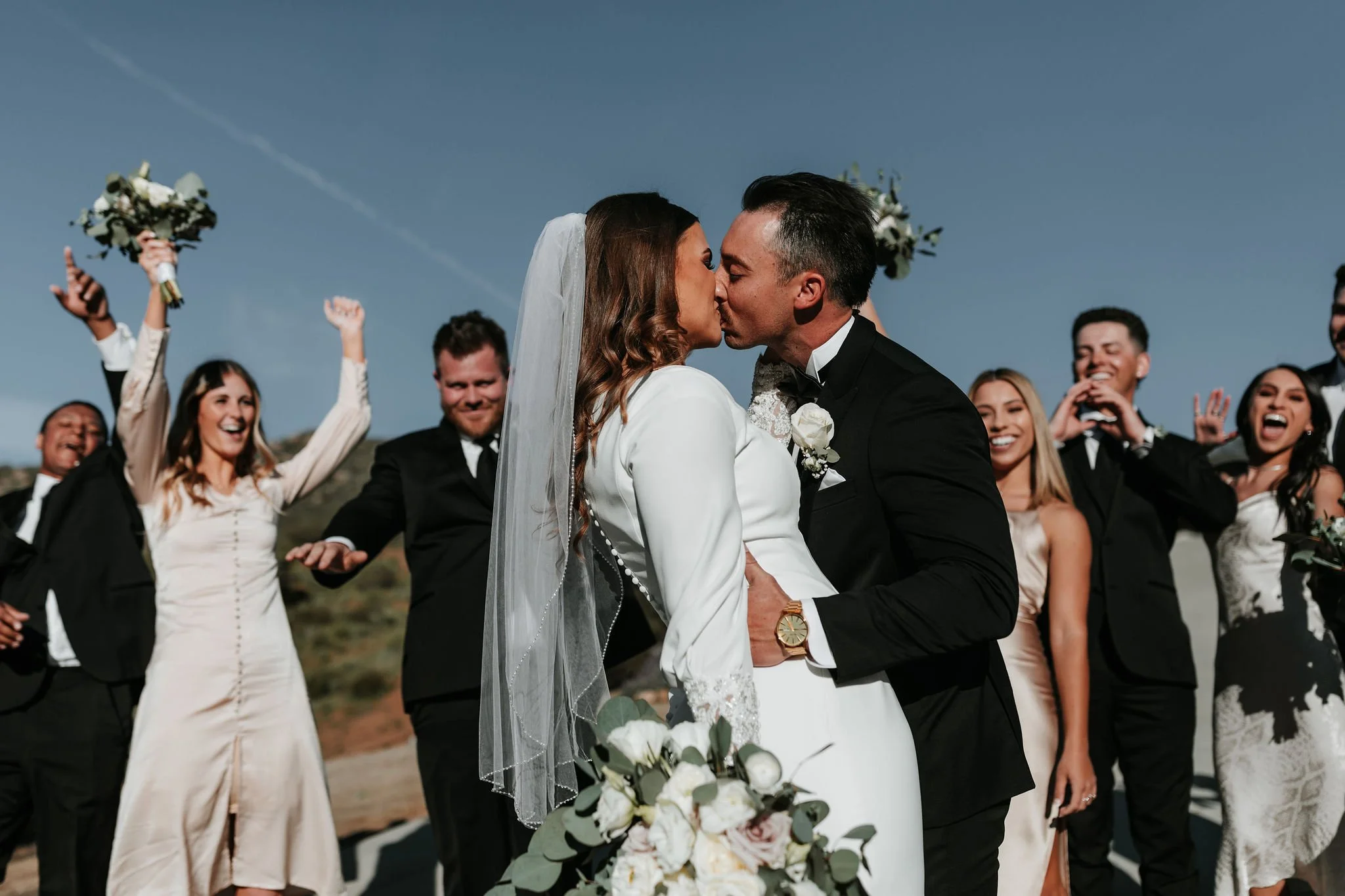 A bride and groom kiss during their outdoor wedding ceremony, surrounded by cheering guests on a clear day.