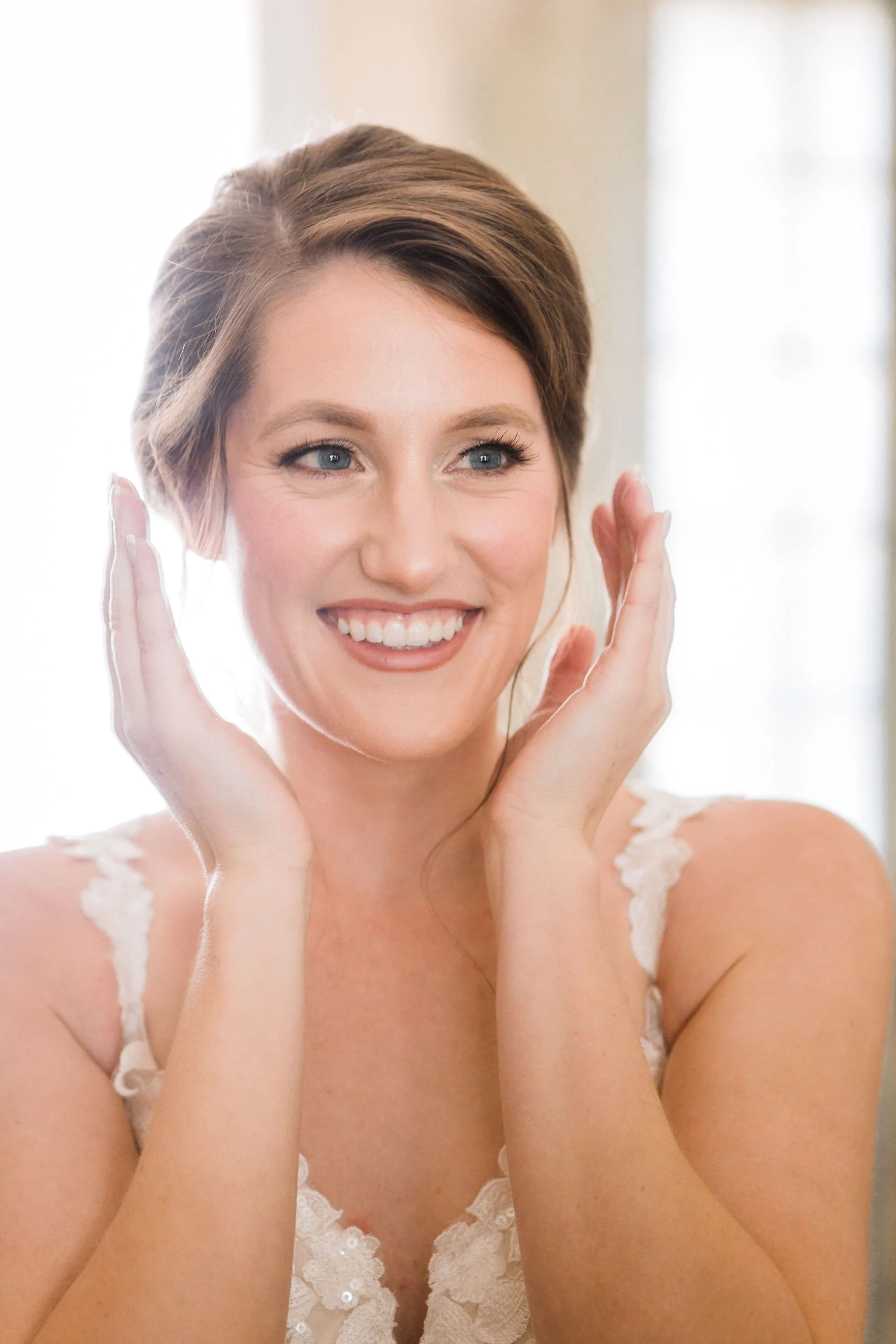 A smiling woman with light brown hair and blue eyes, holding her hands near her face, wearing a white lace top, indoors with soft natural light.