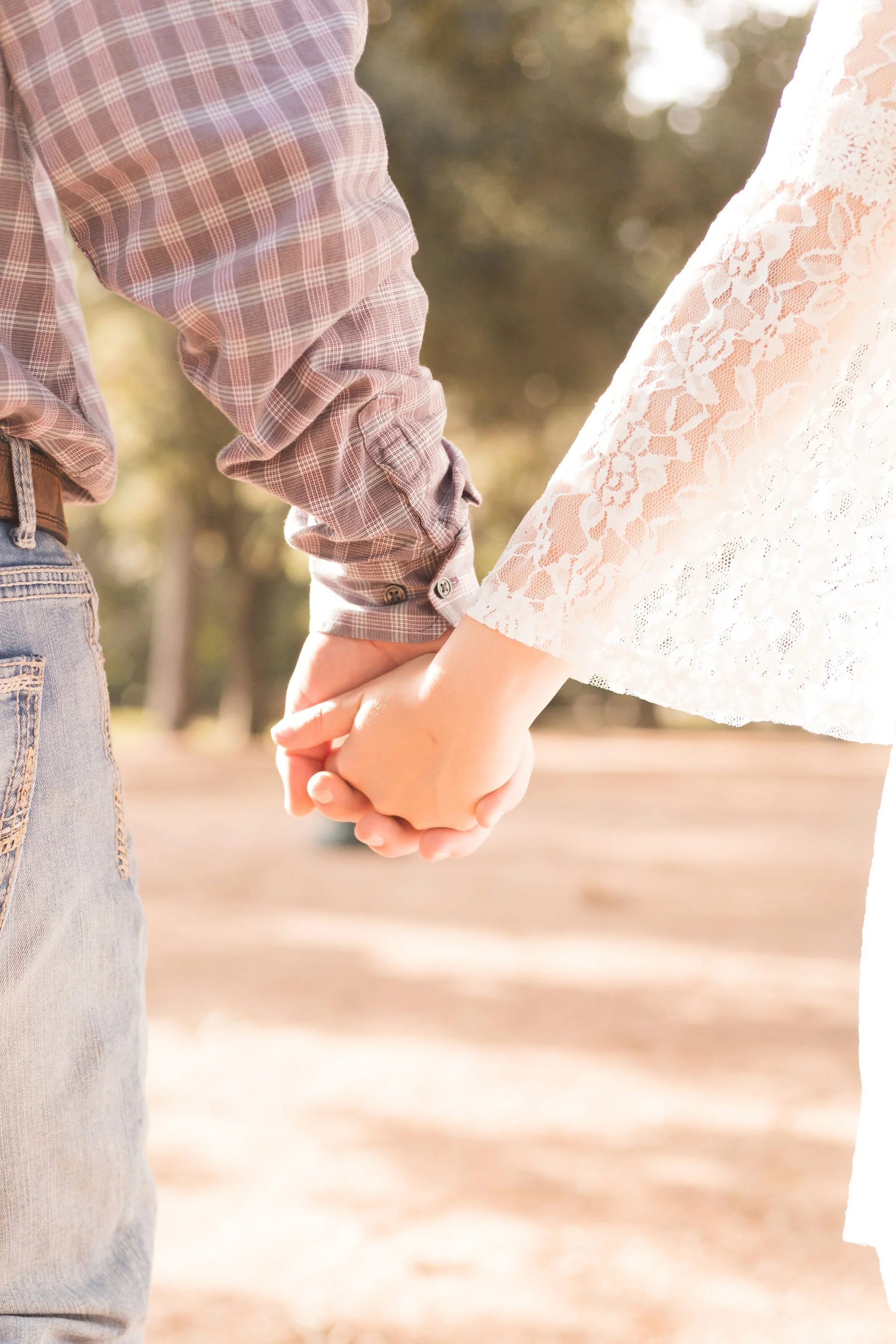 Close-up of a couple holding hands outdoors, woman wearing a white lace dress and man wearing a plaid shirt and jeans.