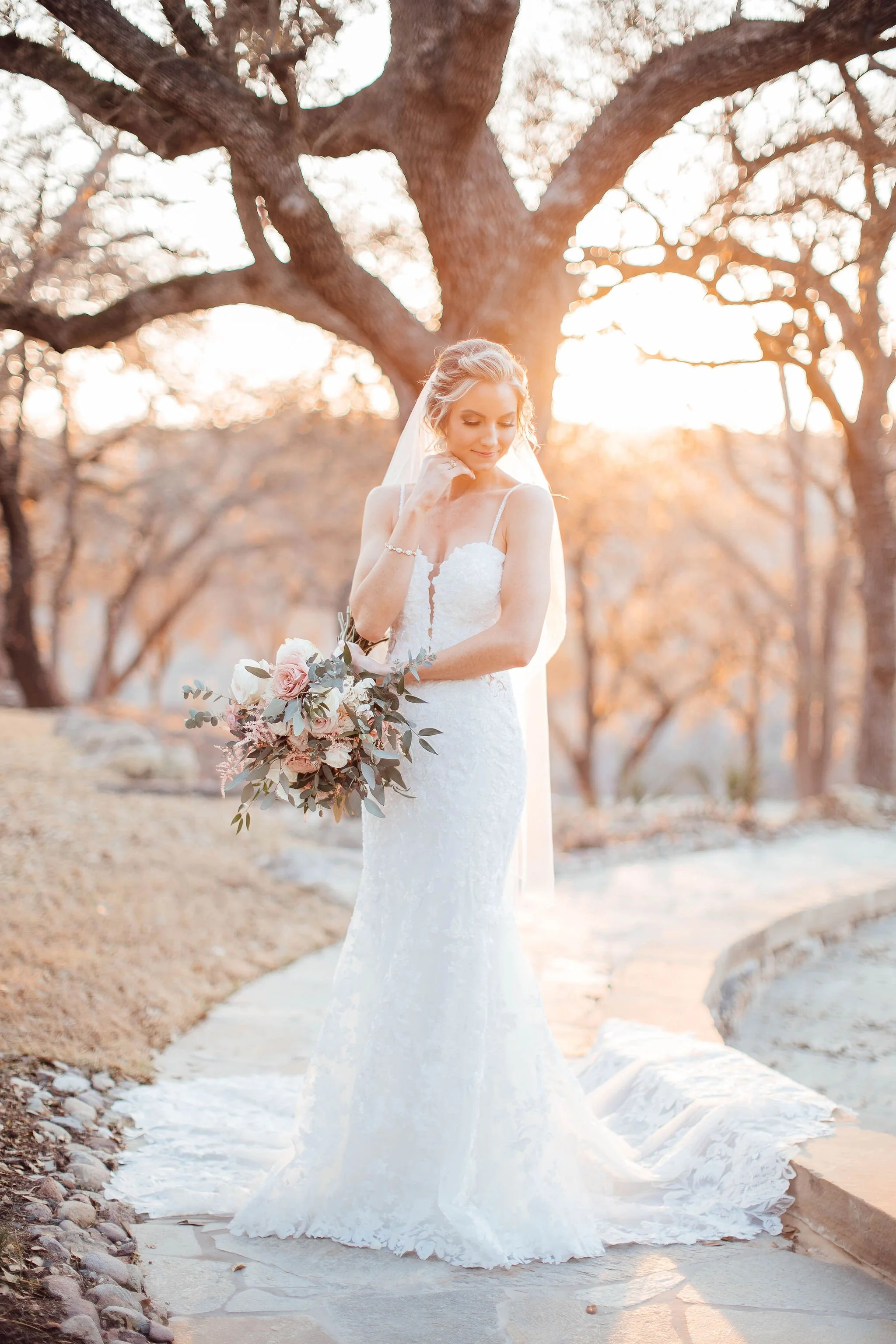A bride wearing a fitted white lace wedding dress stands outdoors on a stone pathway, holding a bouquet of pink and white flowers with greenery. She has blonde hair styled in an elegant updo and wears a sheer veil. The background features leafless tr
