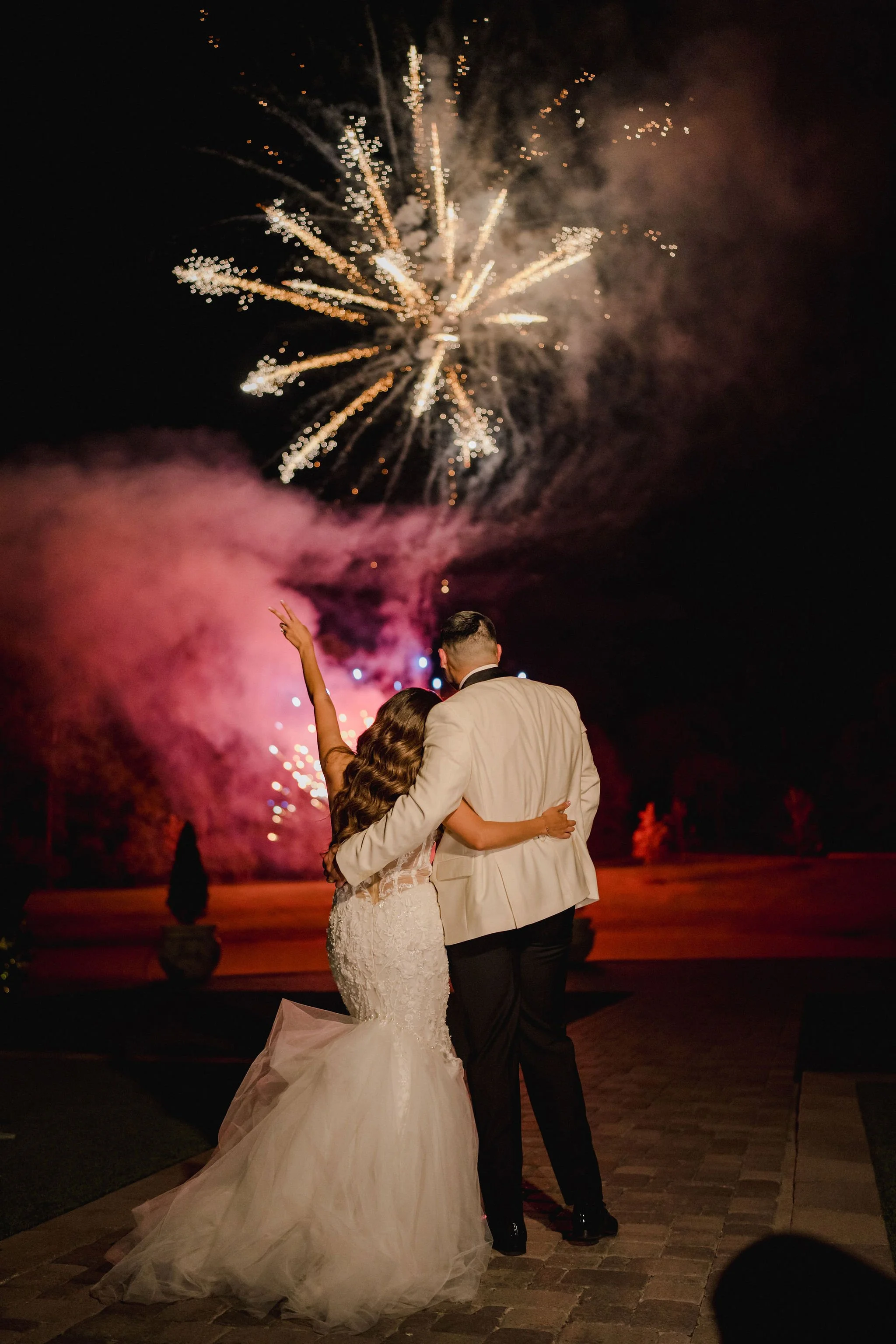 A bride and groom embrace as they watch fireworks light up the night sky during their wedding celebration. The bride is wearing a white wedding gown, and the groom is in a white tuxedo jacket. Fireworks explode in various colors above them, creating 