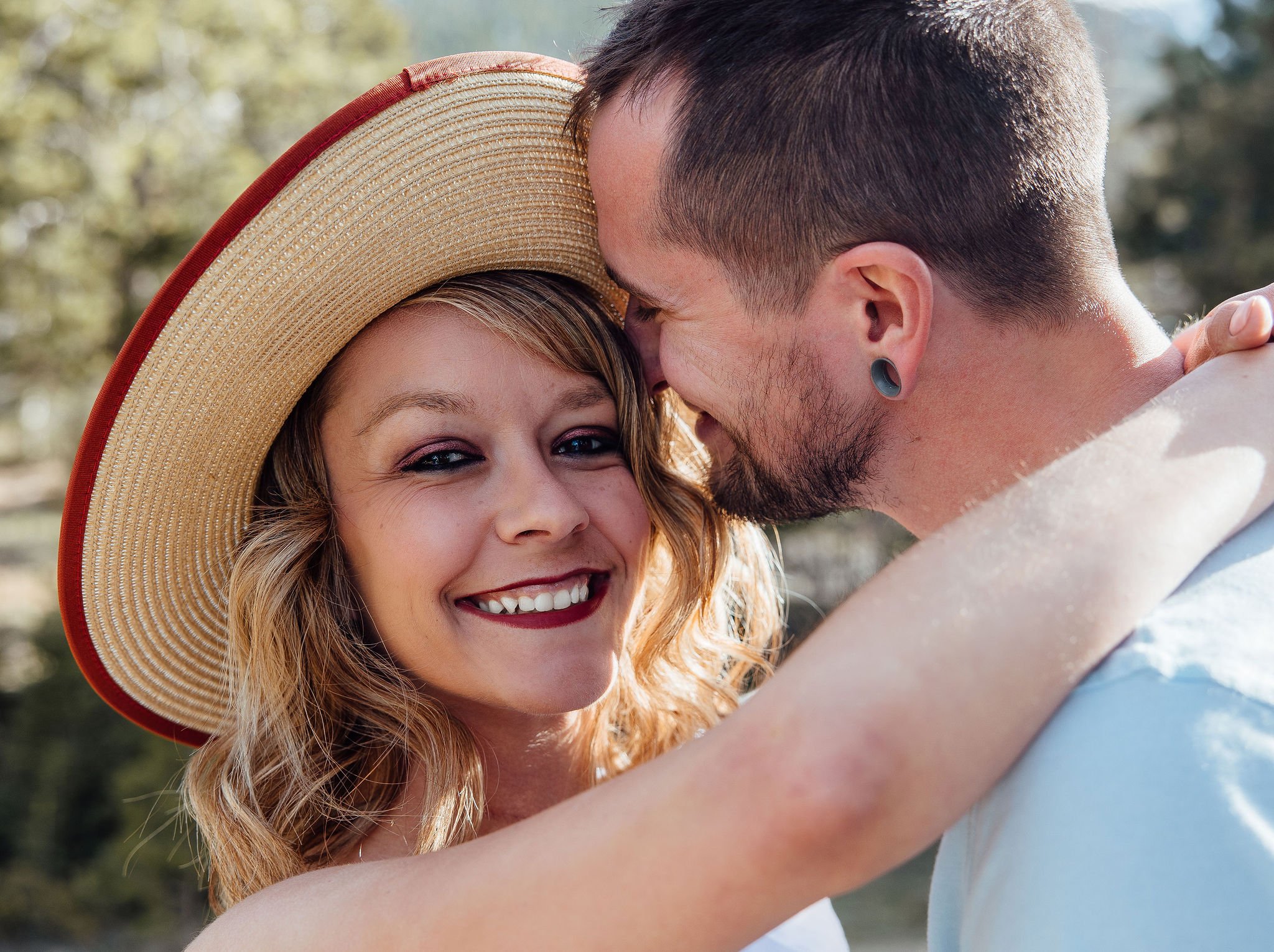 A smiling woman with wavy hair wearing a large straw hat hugging a man with a beard and short hair, both outdoors in sunlight.