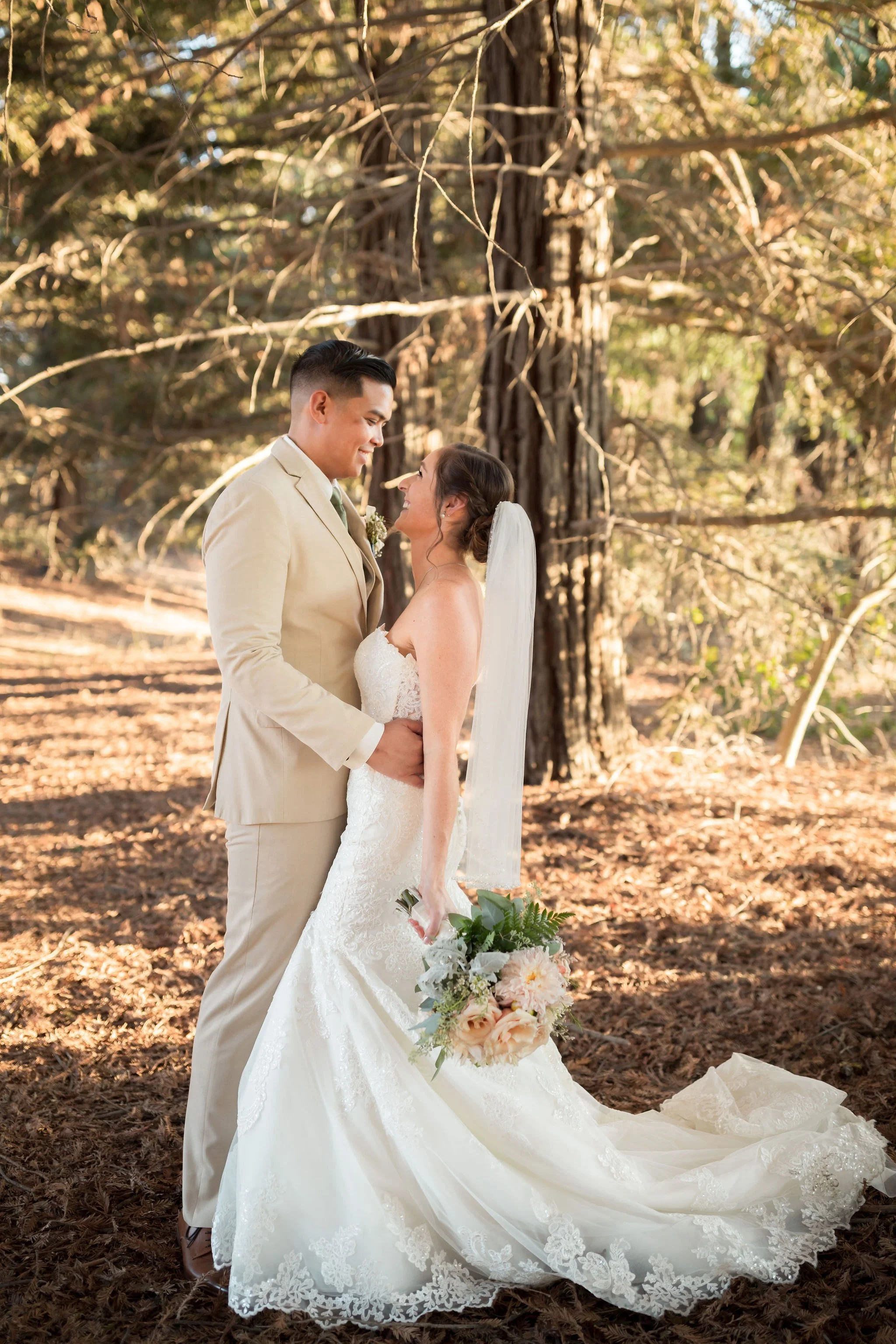 A bride and groom in wedding attire standing close in a forest, looking at each other and smiling. The bride is holding a bouquet of flowers, and there are tall trees and sunlight filtering through the branches.