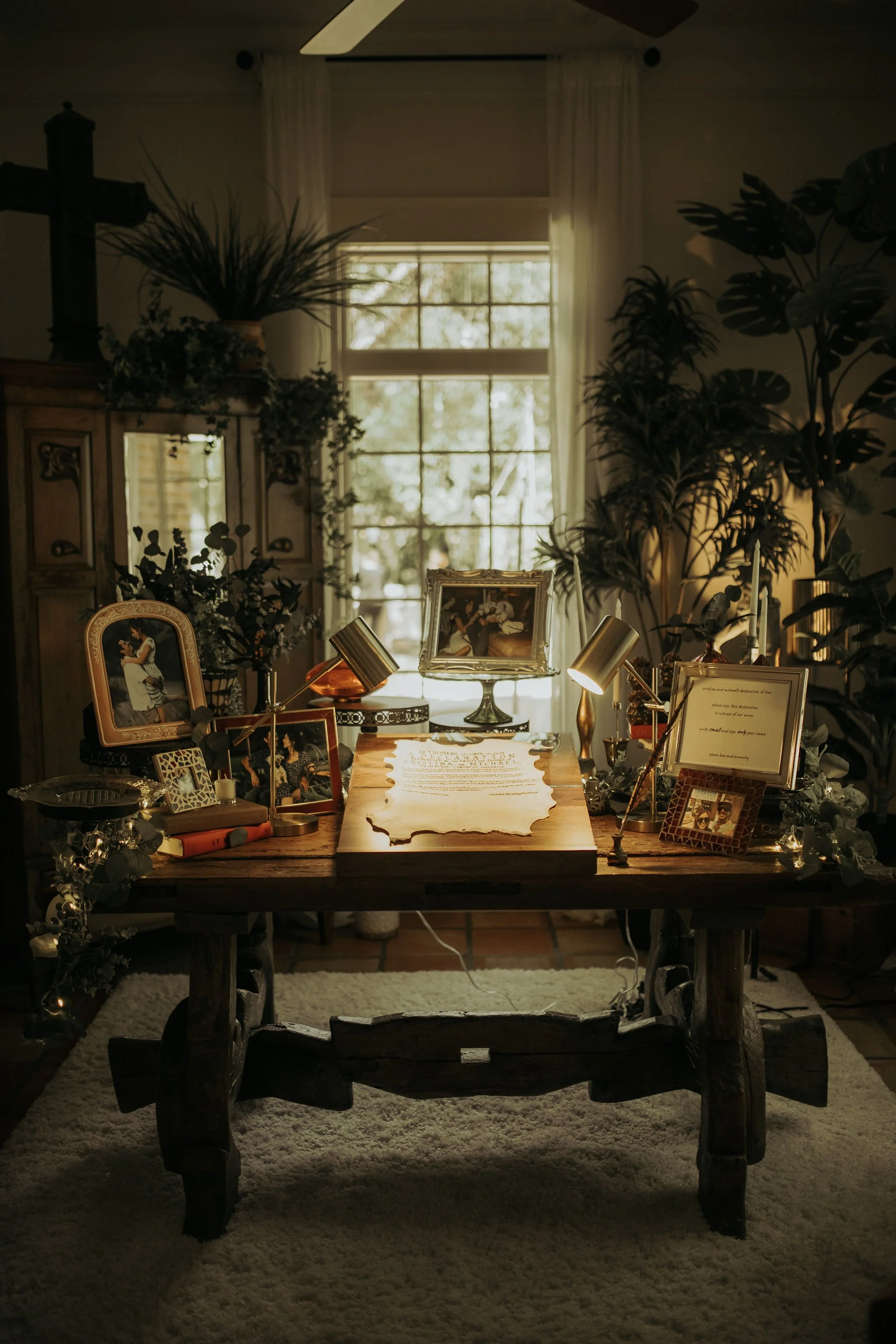 A rustic wooden table decorated with framed photographs, a framed handwritten note, and vintage-style lamps, situated in a room with large window and greenery outside, creating a cozy and vintage ambiance.