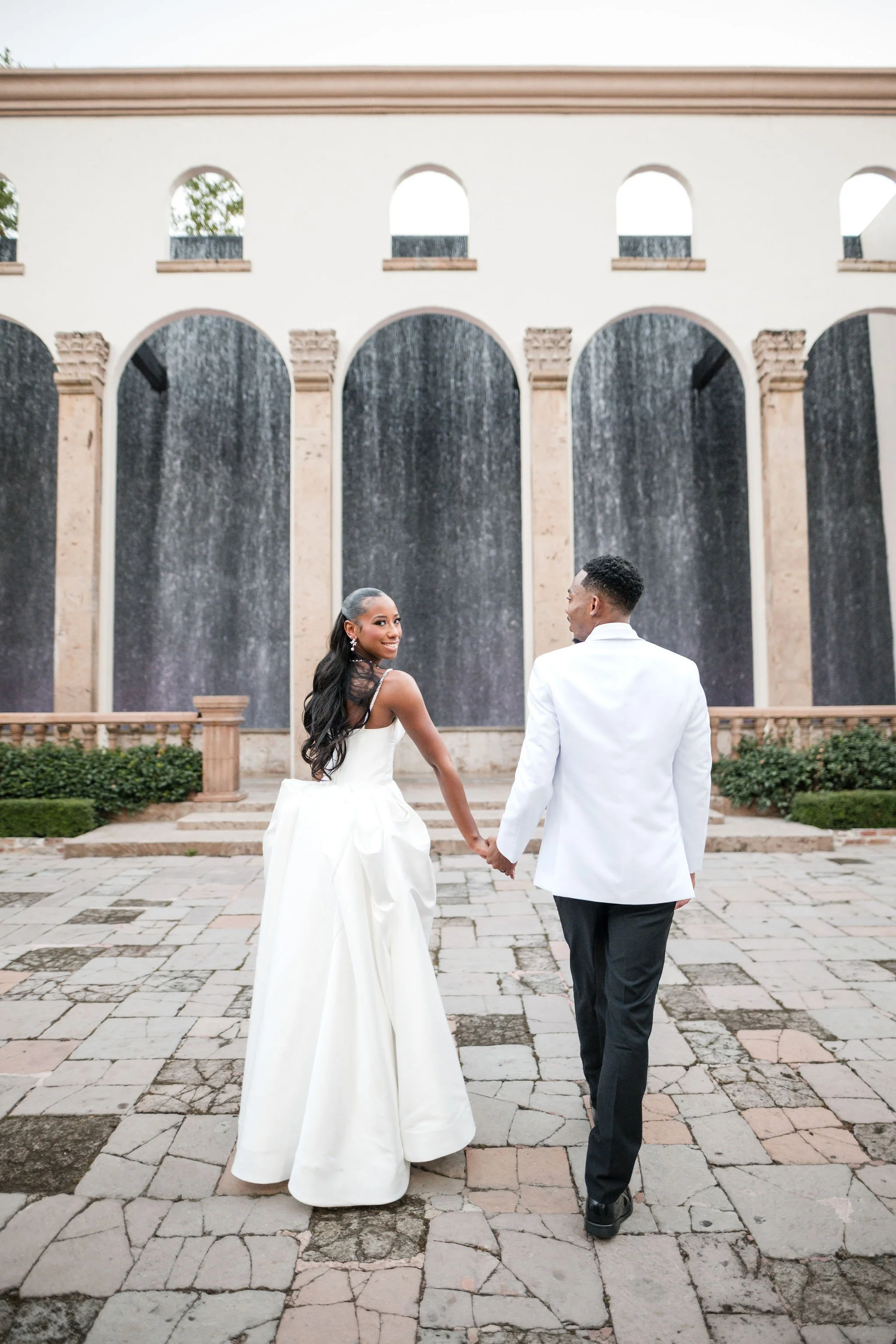 A bride and groom holding hands, walking hand in hand in front of a large decorative wall with arches and water features, on a stone pathway during their wedding.