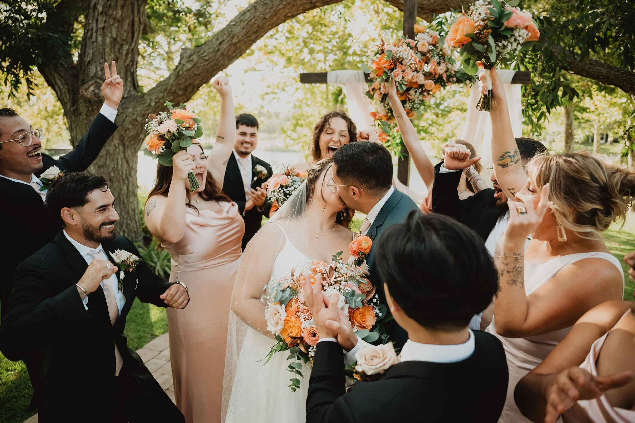 A wedding celebration with a bride and groom kissing surrounded by friends and family outdoors, holding bouquets, smiling, and raising hands in celebration under a large tree with a floral arch in the background.