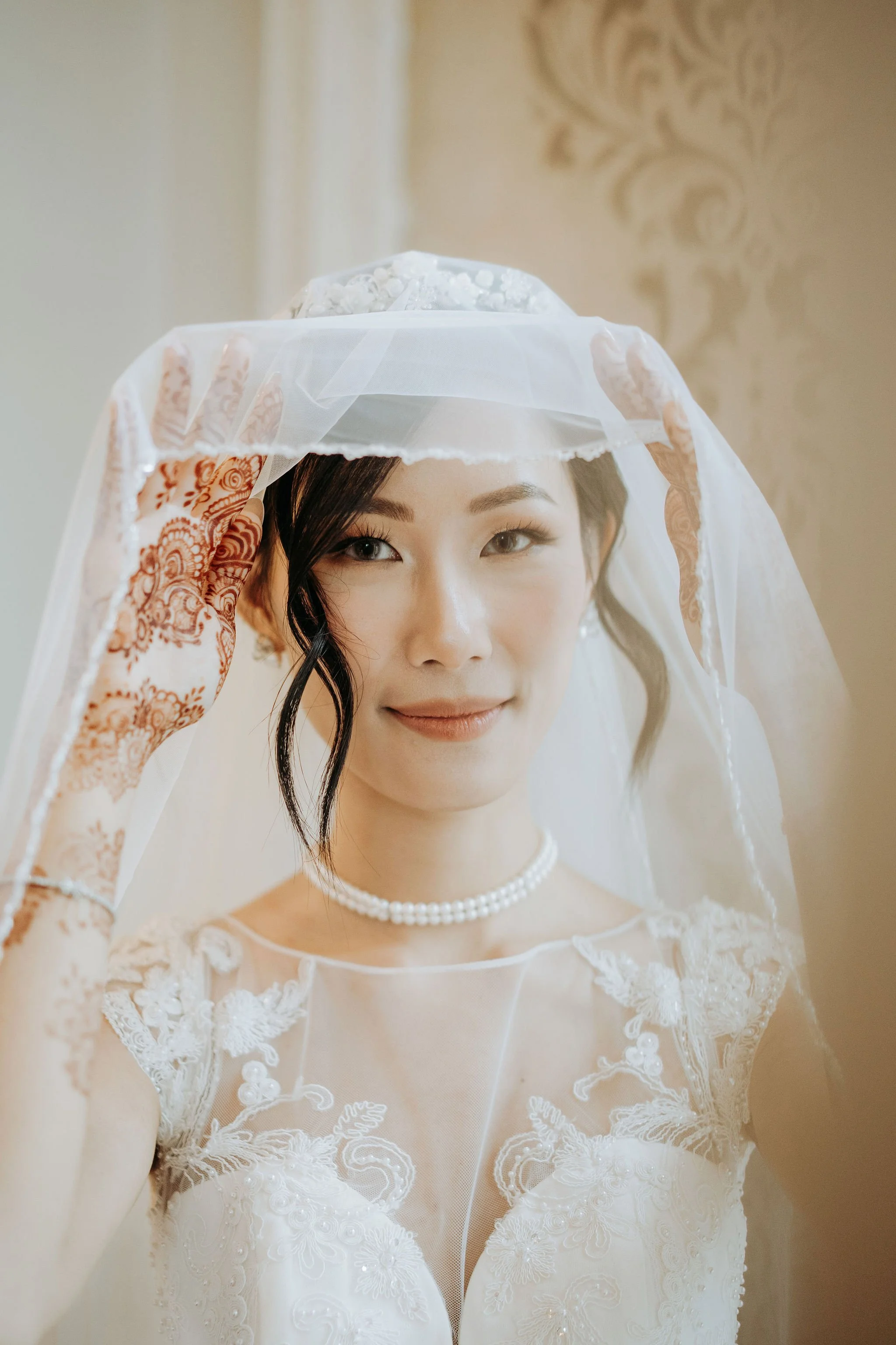 A bride with a pearl necklace adjusting her veil on her head, smiling softly, in a wedding dress with lace details.