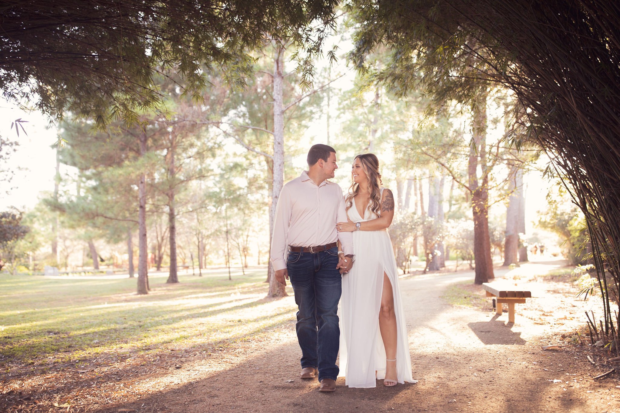A couple holding hands and walking through a sunlit park, smiling at each other. The woman is wearing a white dress with a thigh-high slit, and the man is dressed in a white shirt and jeans. They are surrounded by trees and sunlight.