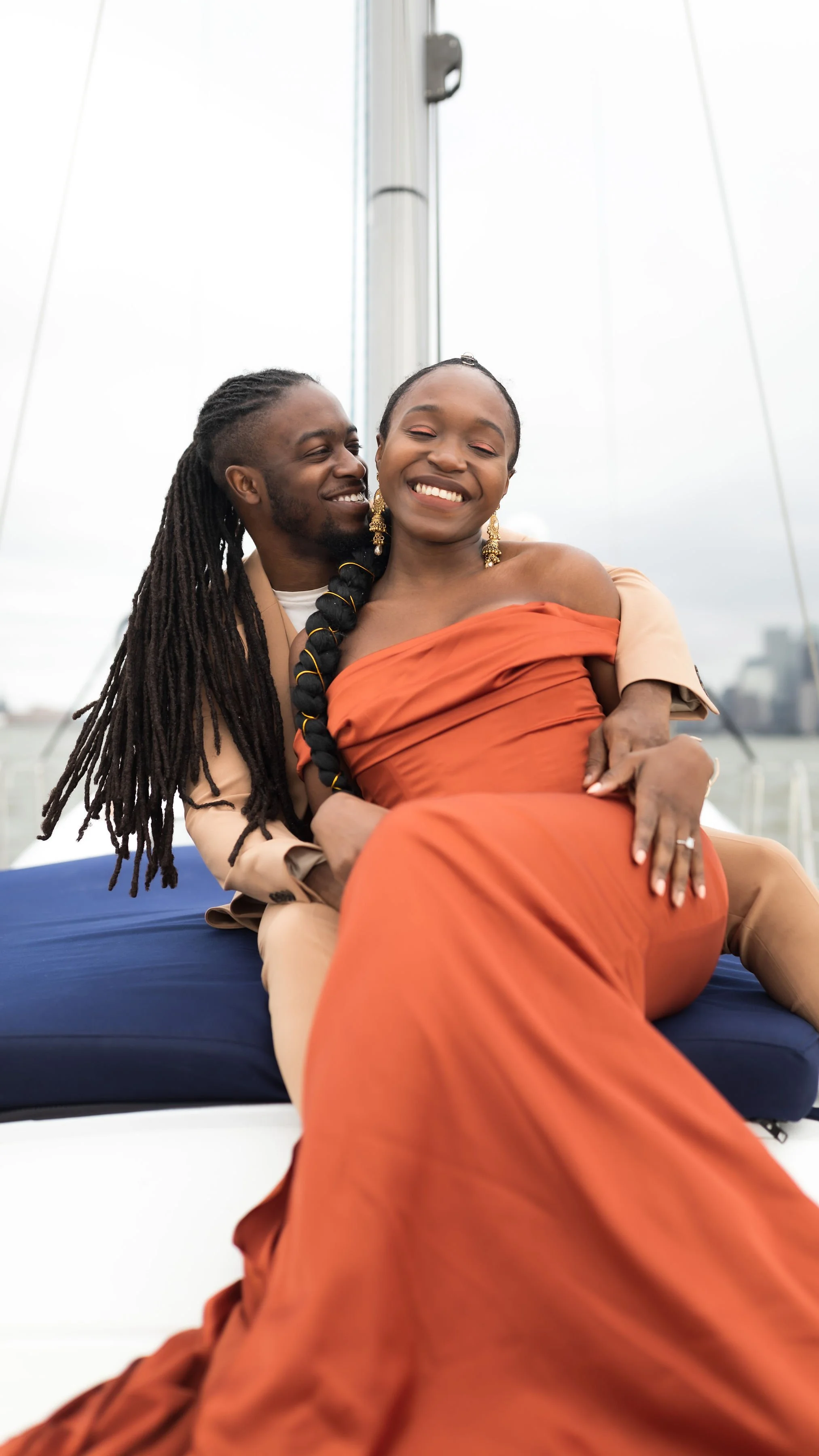 A smiling Black woman in an orange off-shoulder dress sitting on a boat with a man in beige suit, with gray sky and harbor in the background.
