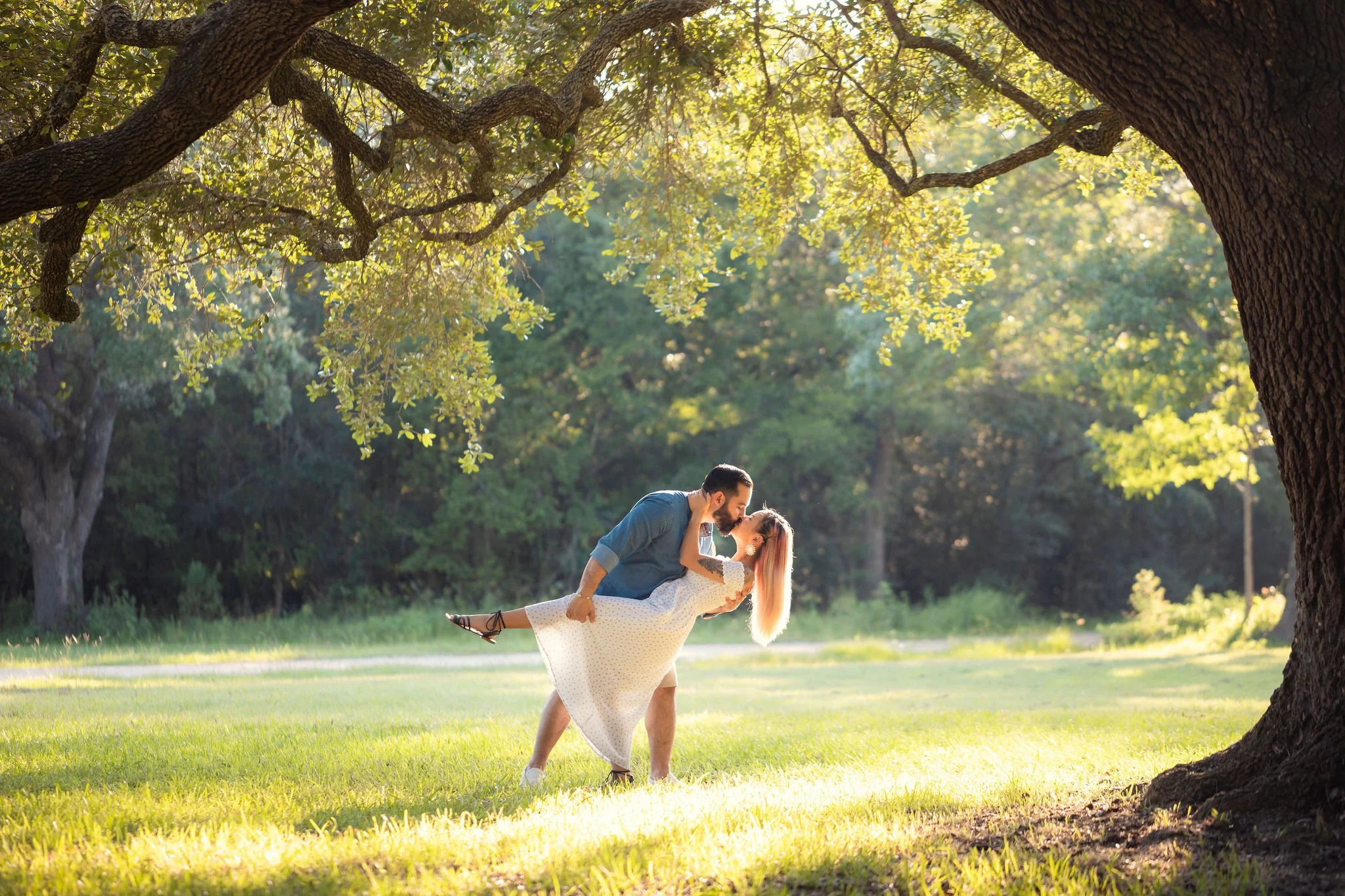 A man and woman dancing and sharing a kiss outdoors in a park surrounded by trees and green grass, with sunlight filtering through the leaves.