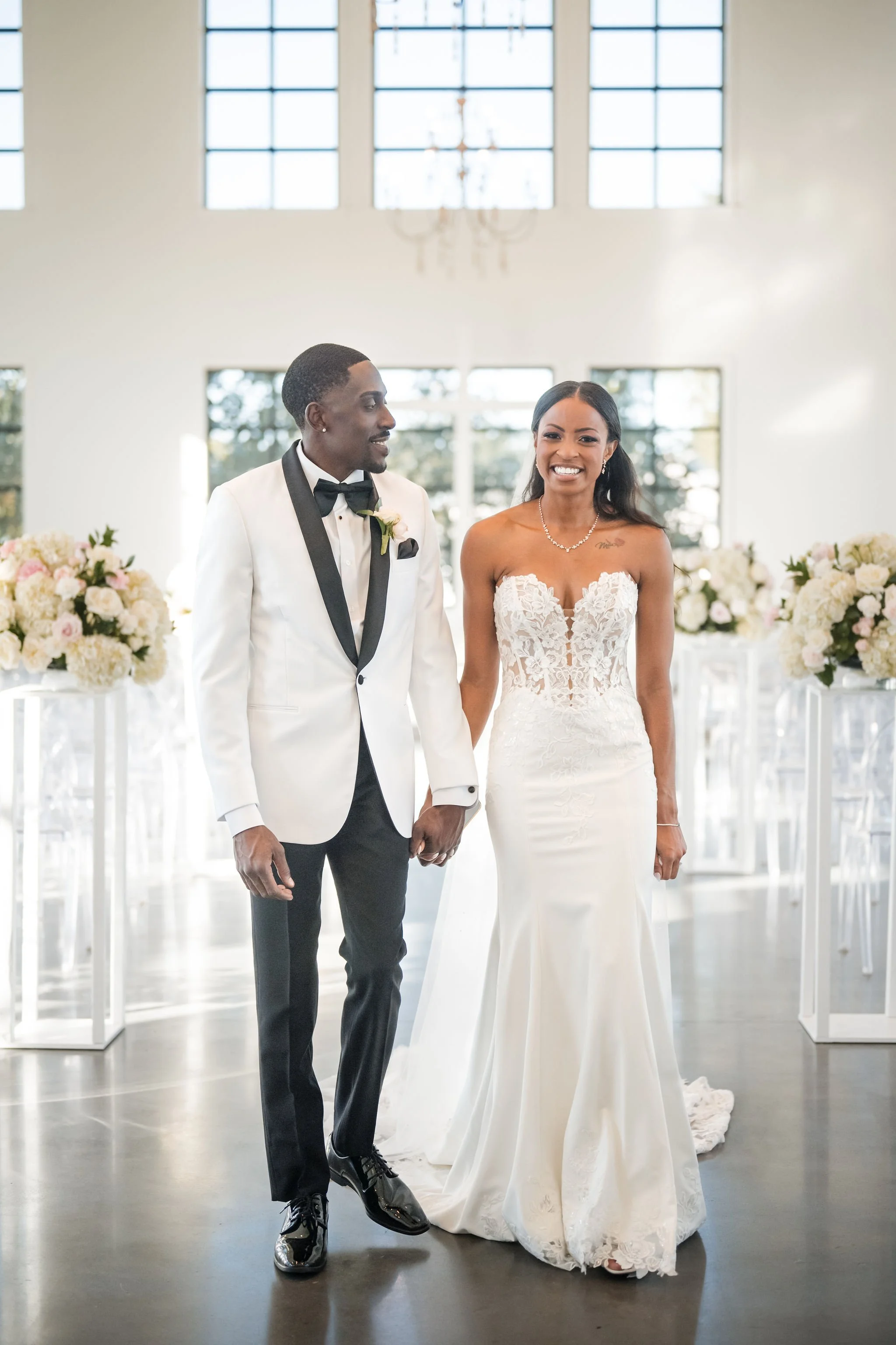 A bride and groom holding hands and smiling during their wedding ceremony in a bright indoor venue decorated with flower arrangements.