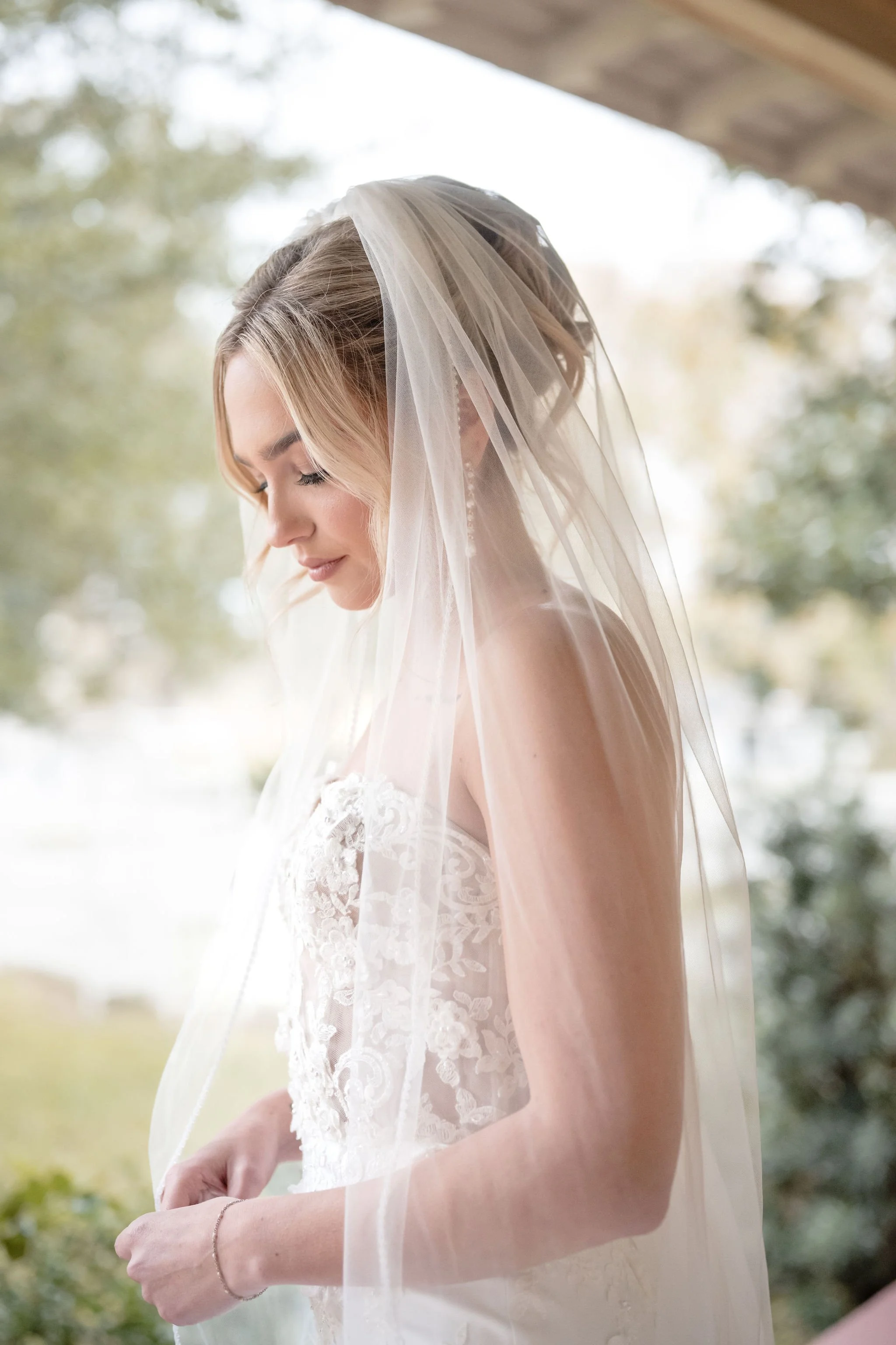 A bride wearing a lace wedding dress and a veil, standing outdoors with her eyes closed and a gentle smile.